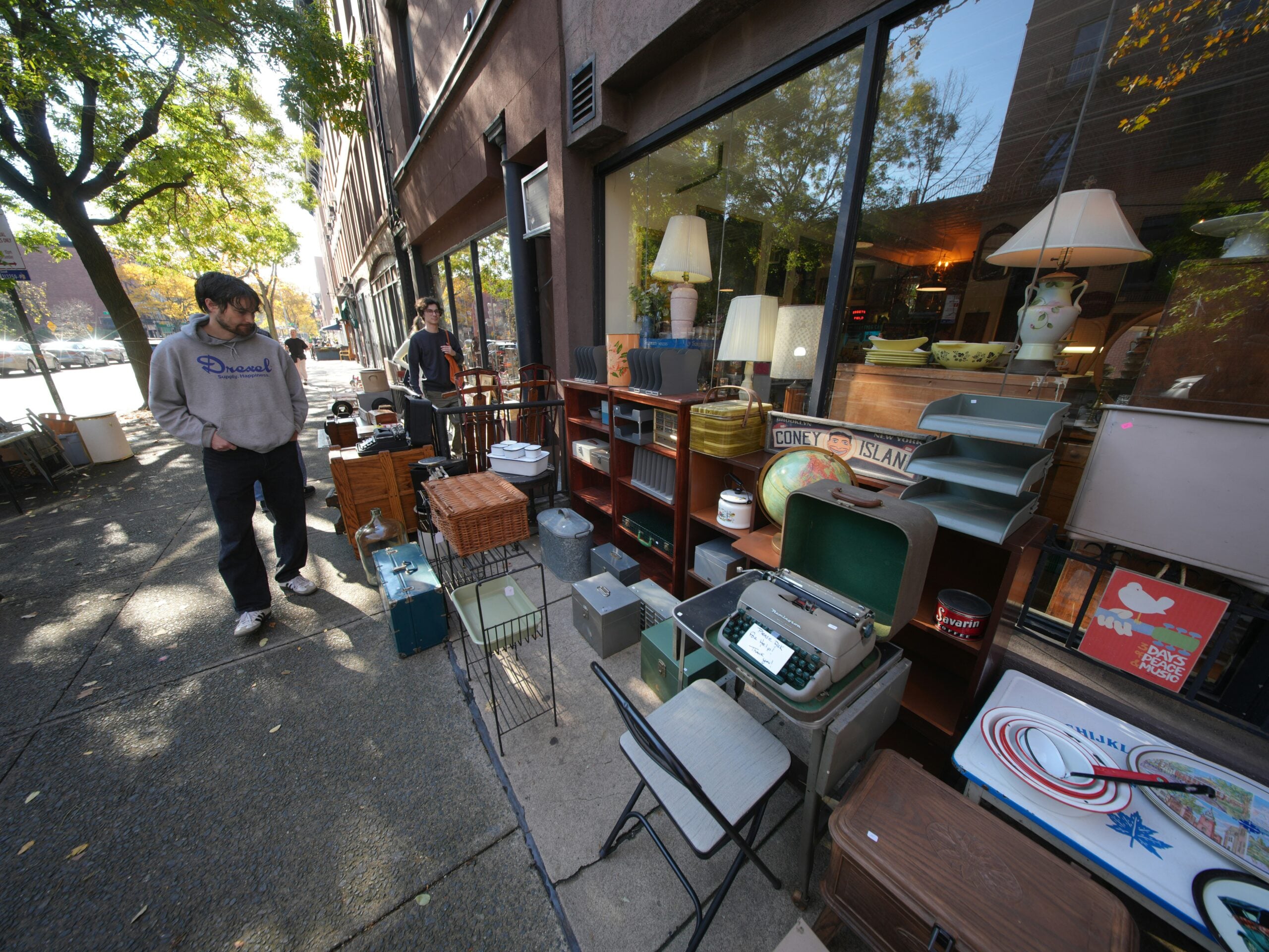 Two people walk past a vintage shop with shelves, lamps, baskets, and various retro items displayed on the sidewalk outside the storefront. Trees line the street on a sunny day.