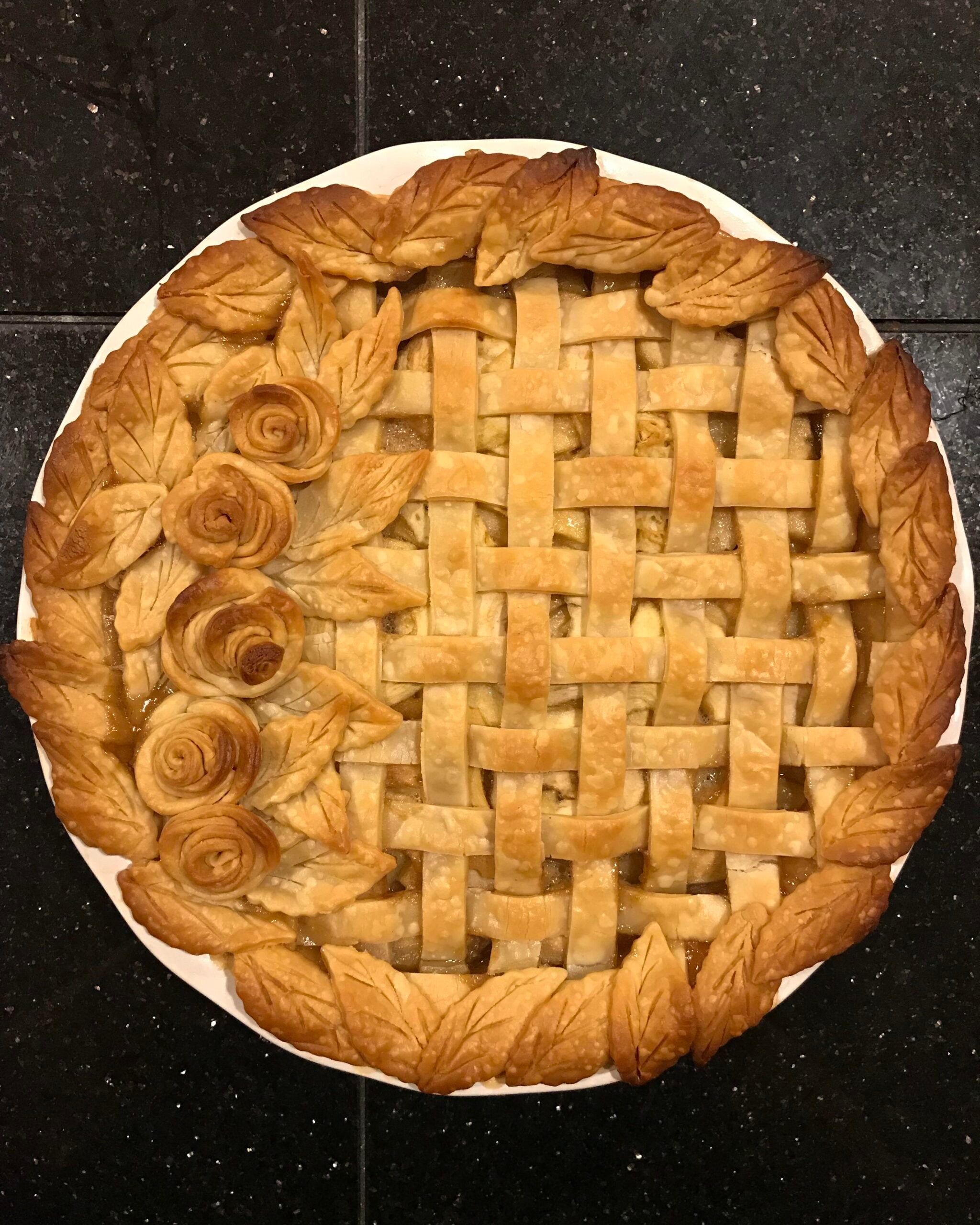 A pie with a lattice crust, decorated with leaf-shaped and rose-shaped pastry designs on a black tiled surface.