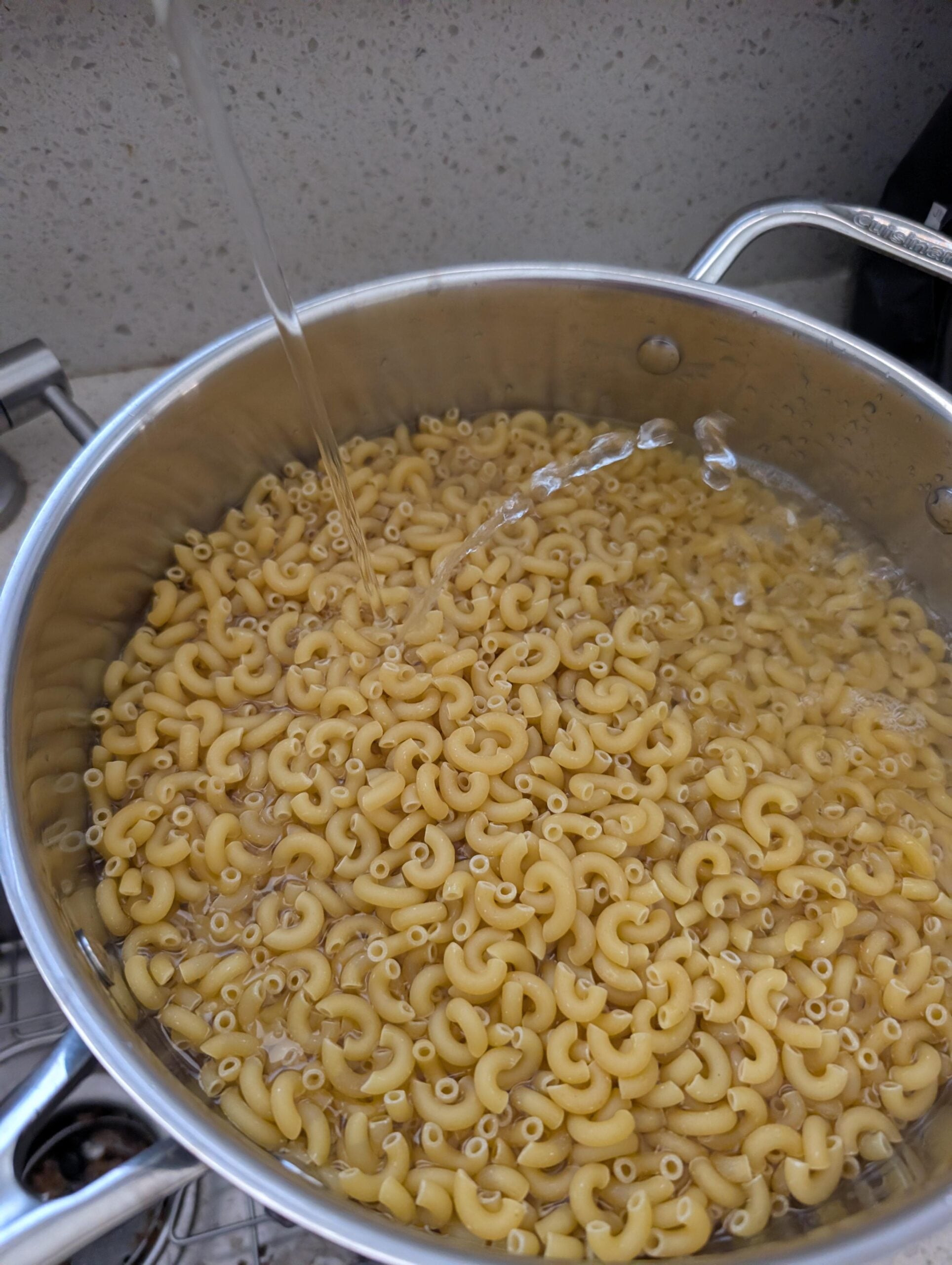 A pot filled with uncooked elbow macaroni is being filled with water from a tap, preparing the pasta for boiling.