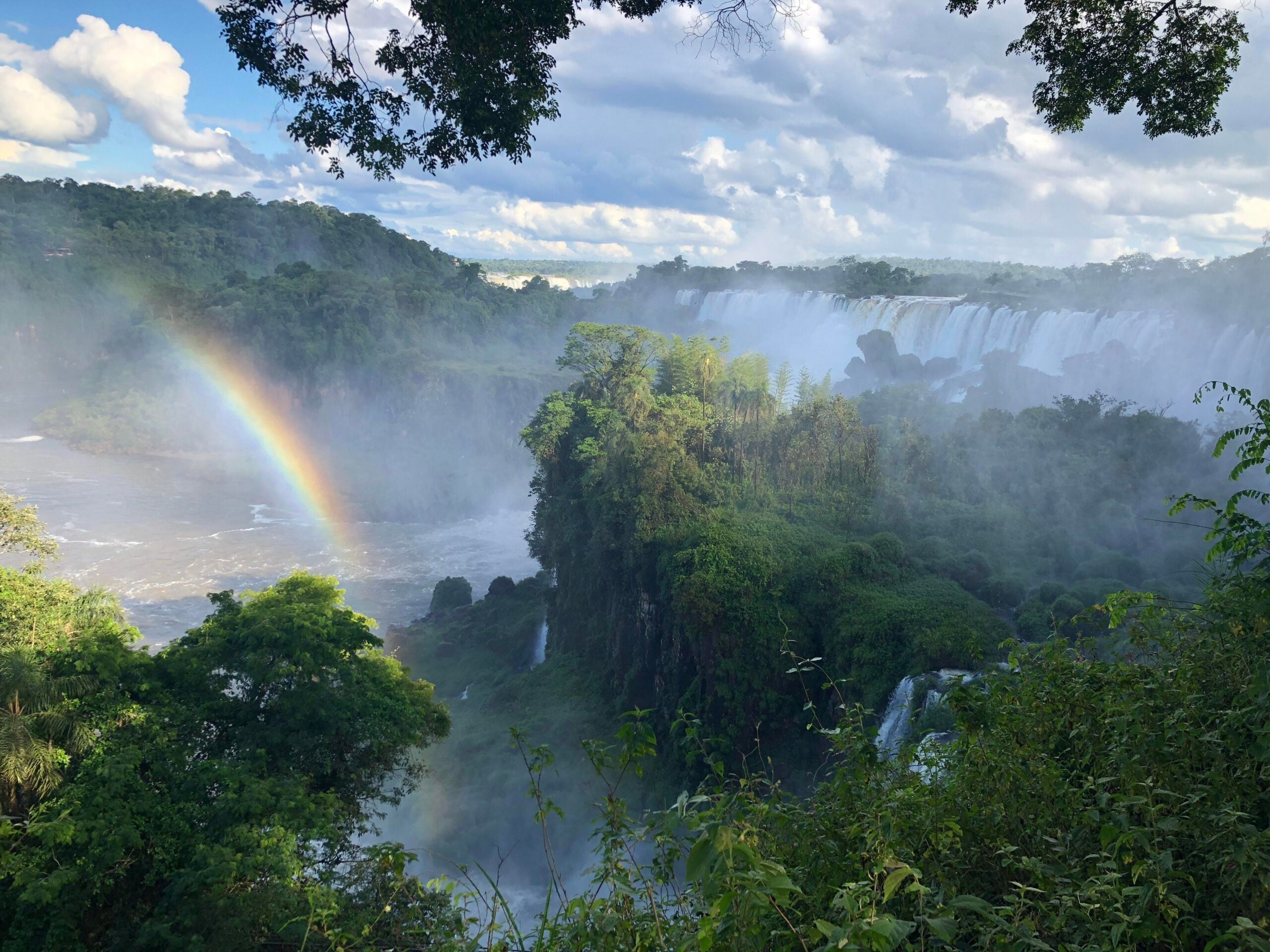 Large waterfalls cascade through dense green forest under a cloudy sky, with mist rising around the falls. A vivid rainbow arches over the water in the foreground, adding color to the lush landscape.
