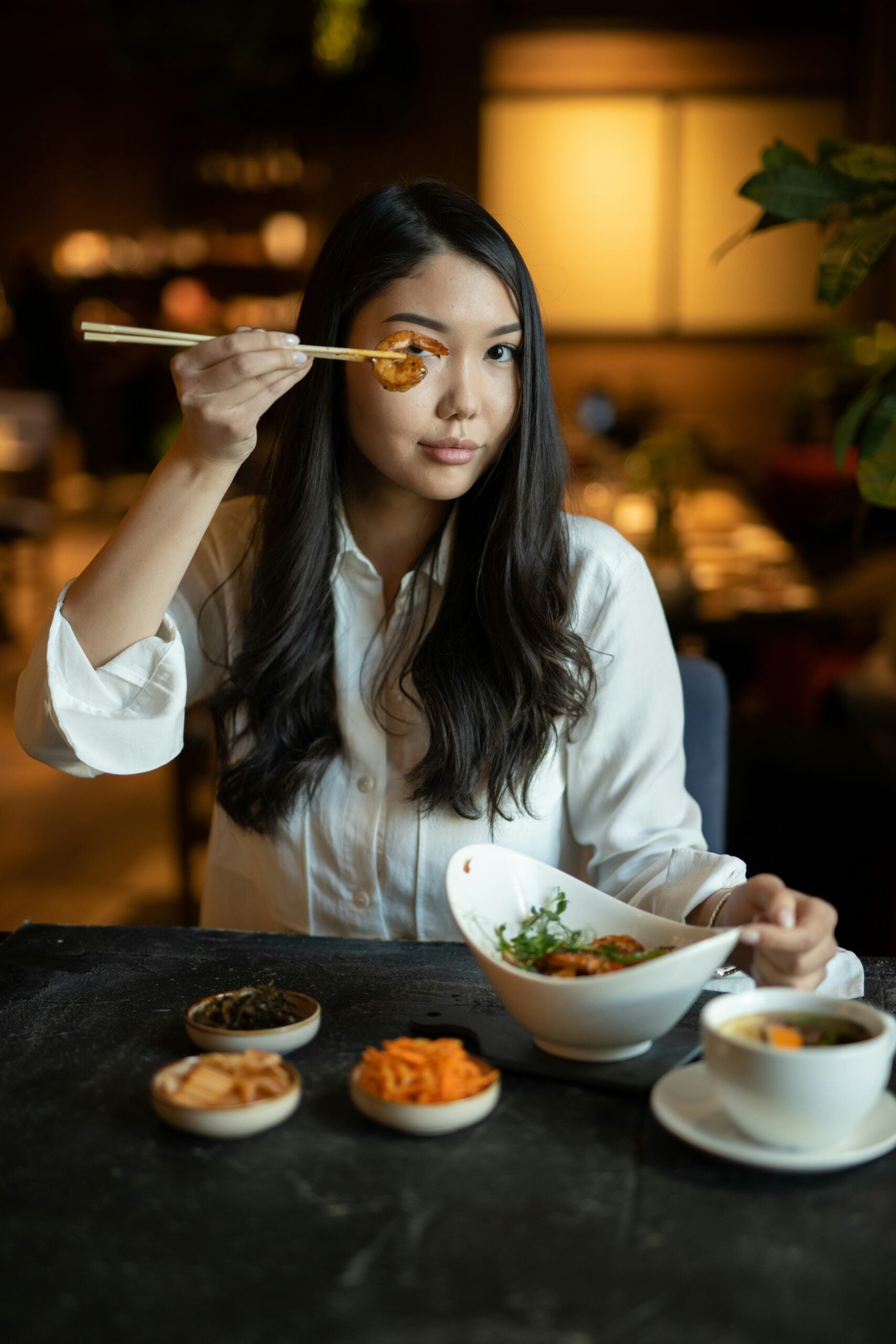 A woman with long dark hair in a white shirt holds food with chopsticks near her eye, sitting at a table with several small dishes and a bowl of noodles in a warmly lit restaurant.