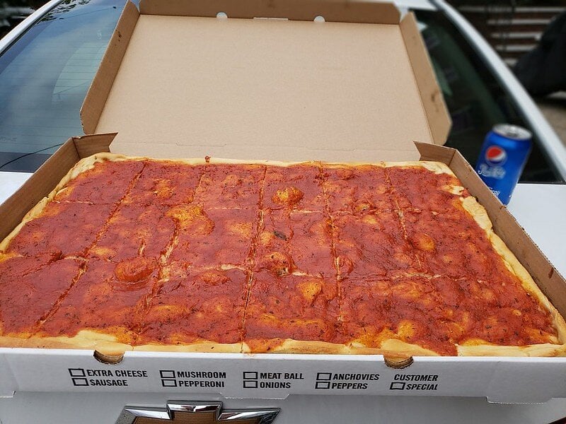 A large rectangular pizza with tomato sauce cut into squares is in an open box on a car hood. A can of Pepsi sits in the background.