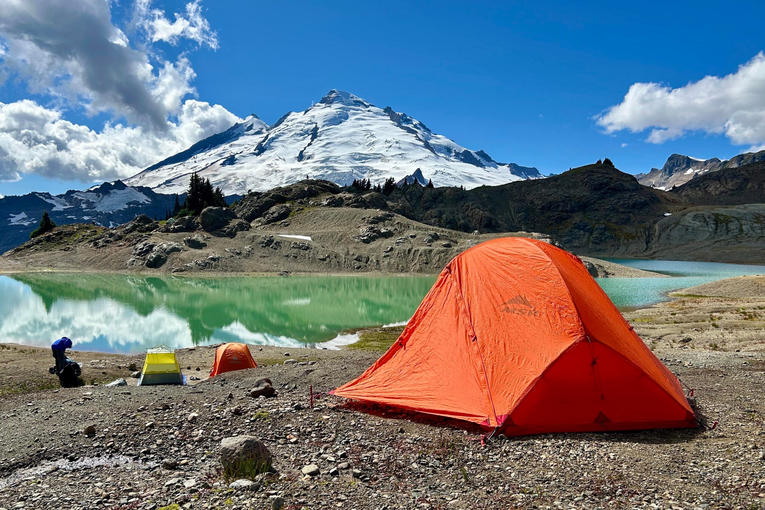 Three tents, including a prominent orange one, are set up by a turquoise lake with a snow-capped mountain in the background under a partly cloudy blue sky.