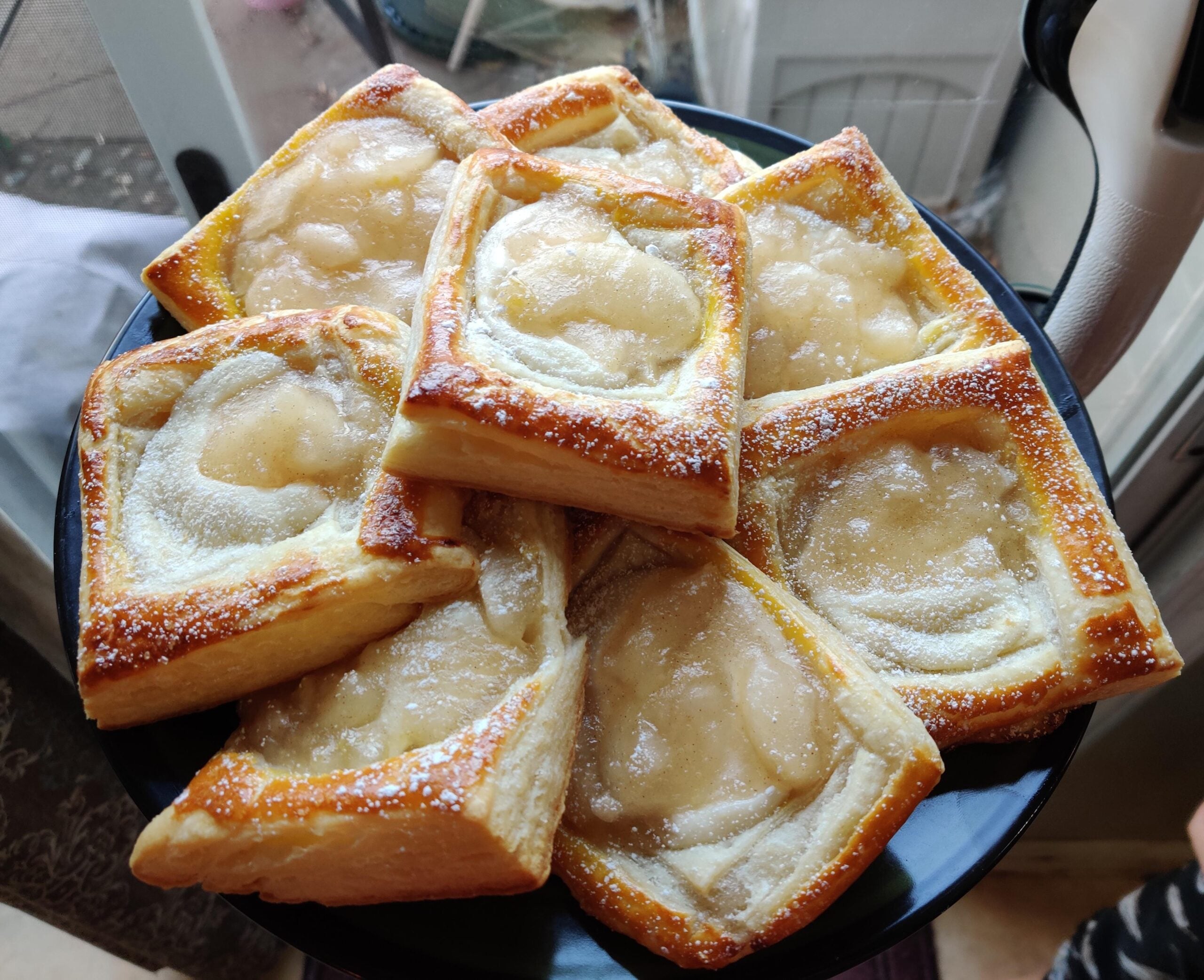 A plate of golden-brown pastries topped with glossy fruit filling, arranged in a stack near a window, with sunlight highlighting their flaky texture.