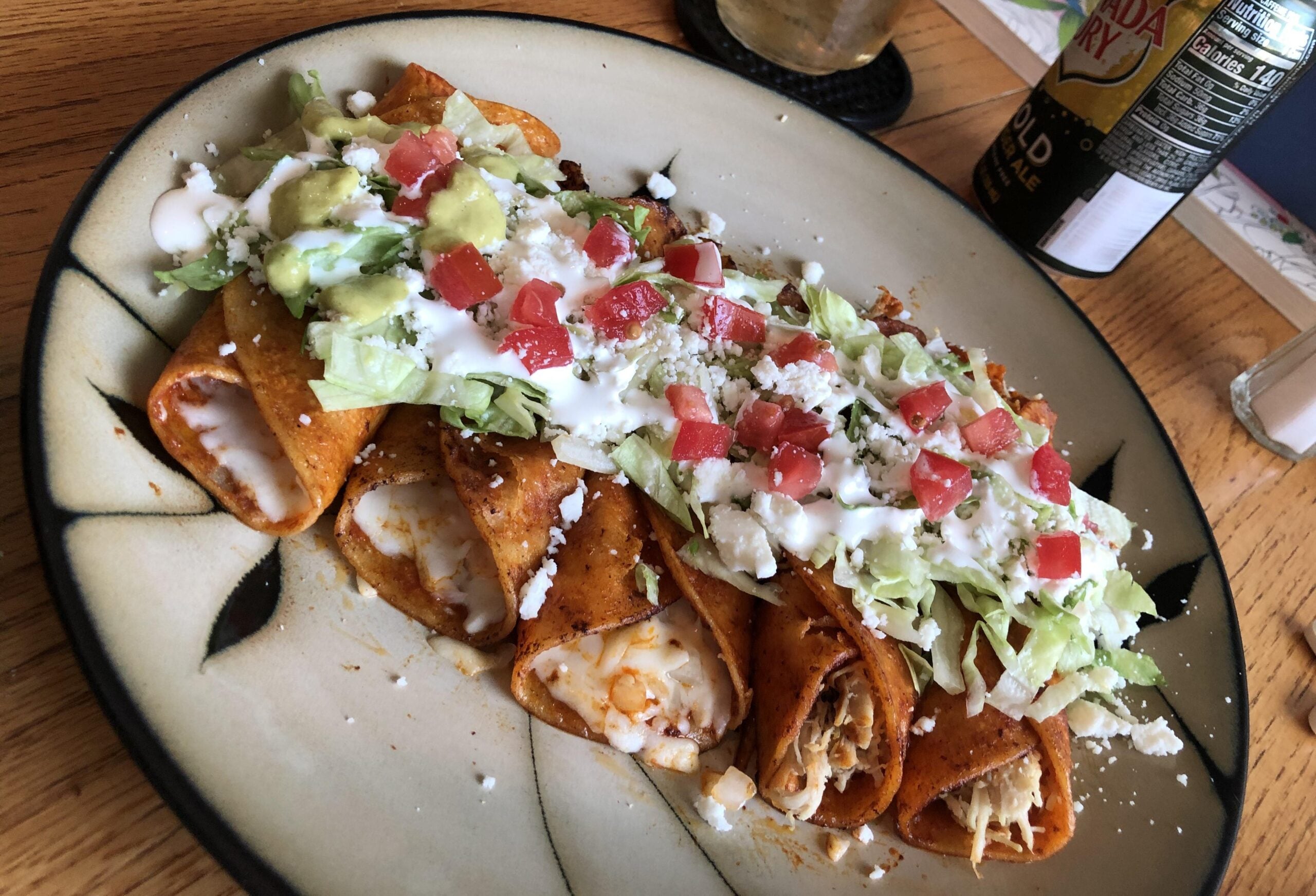 A plate of enchiladas topped with shredded lettuce, diced tomatoes, guacamole, cheese, and a drizzle of sour cream sits on a wooden table beside a drink and a can of hard seltzer.