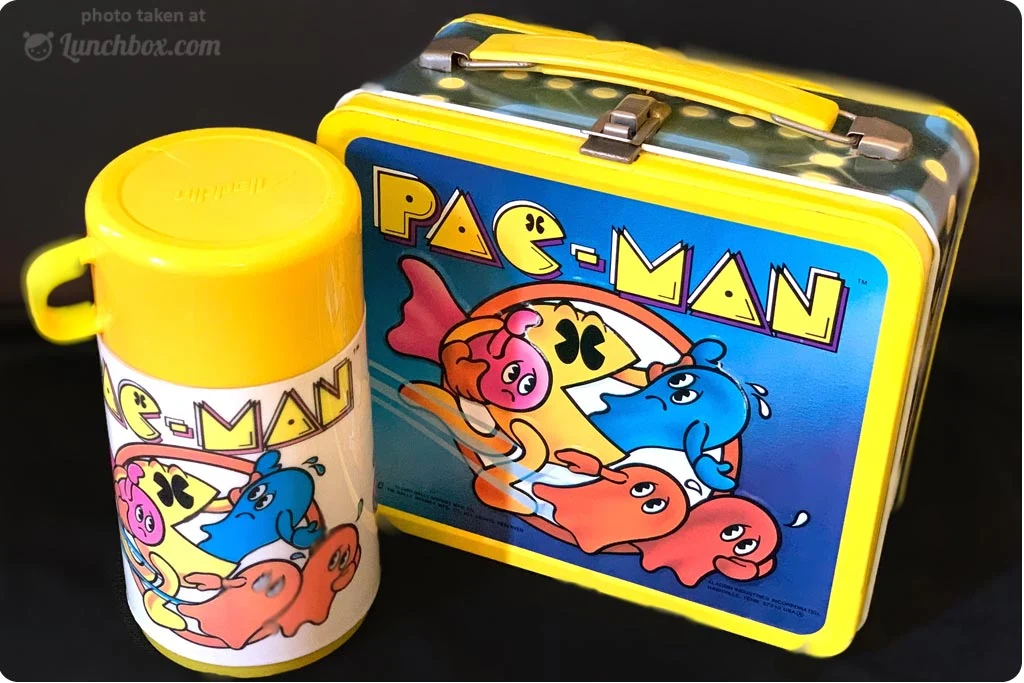 A vintage Pac-Man lunchbox and matching thermos, both decorated with colorful Pac-Man and ghost characters on a bright blue and yellow background.