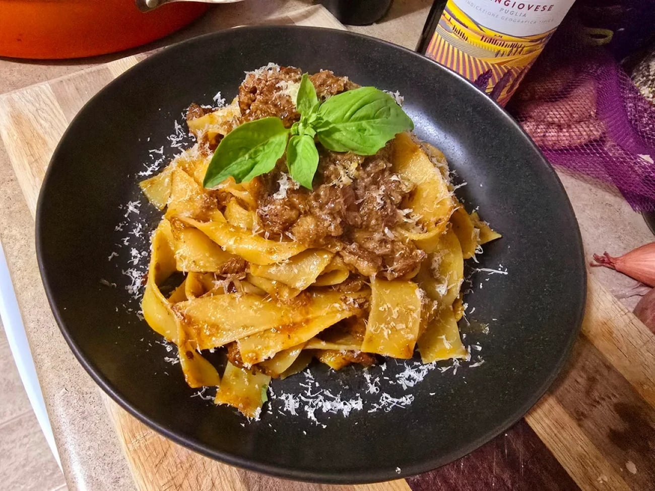 Pappardelle pasta topped with meat sauce, grated cheese, and fresh basil, served on a black plate. Photos of pasta like this often capture the rich textures and inviting colors of the dish, with a bottle of red wine and kitchen items visible in the background.