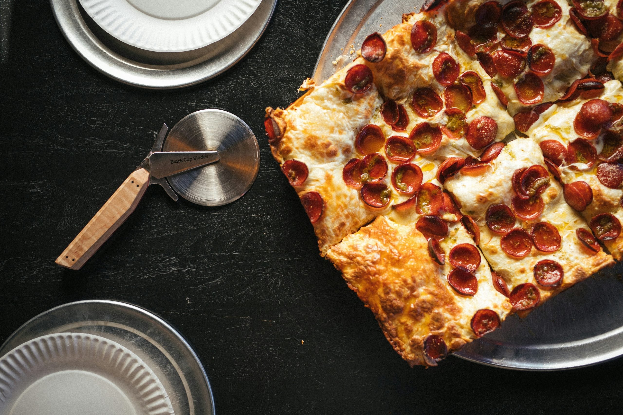 A rectangular pepperoni pizza, one of the classic comfort foods, rests on a metal tray beside a pizza cutter and empty white paper plates on a dark table.