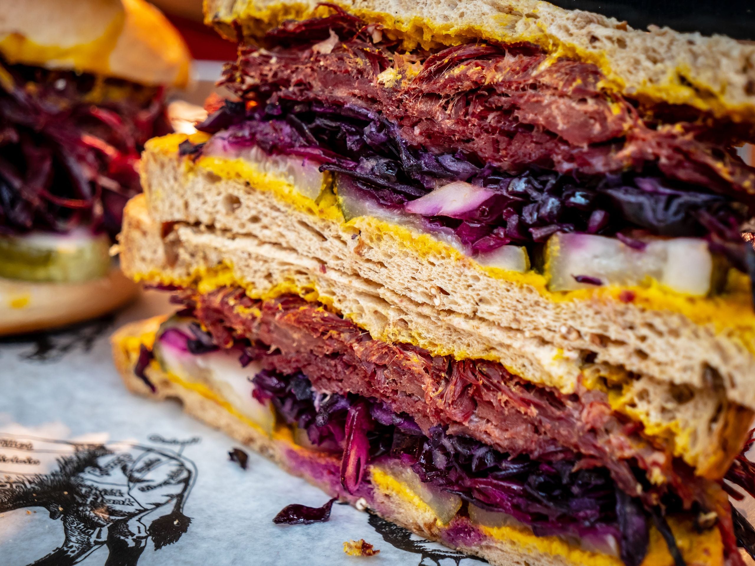 Close-up of a stacked sandwich with layers of corned beef, pickles, shredded red cabbage, and mustard on rye bread—a mouthwatering display of comfort foods. Another sandwich is blurred in the background.