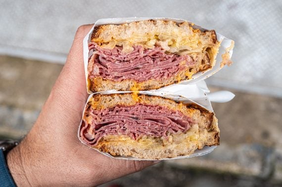 A hand holds a stacked sandwich cut in half, showing layers of pastrami, melted cheese, and cabbage between toasted bread, wrapped in paper.