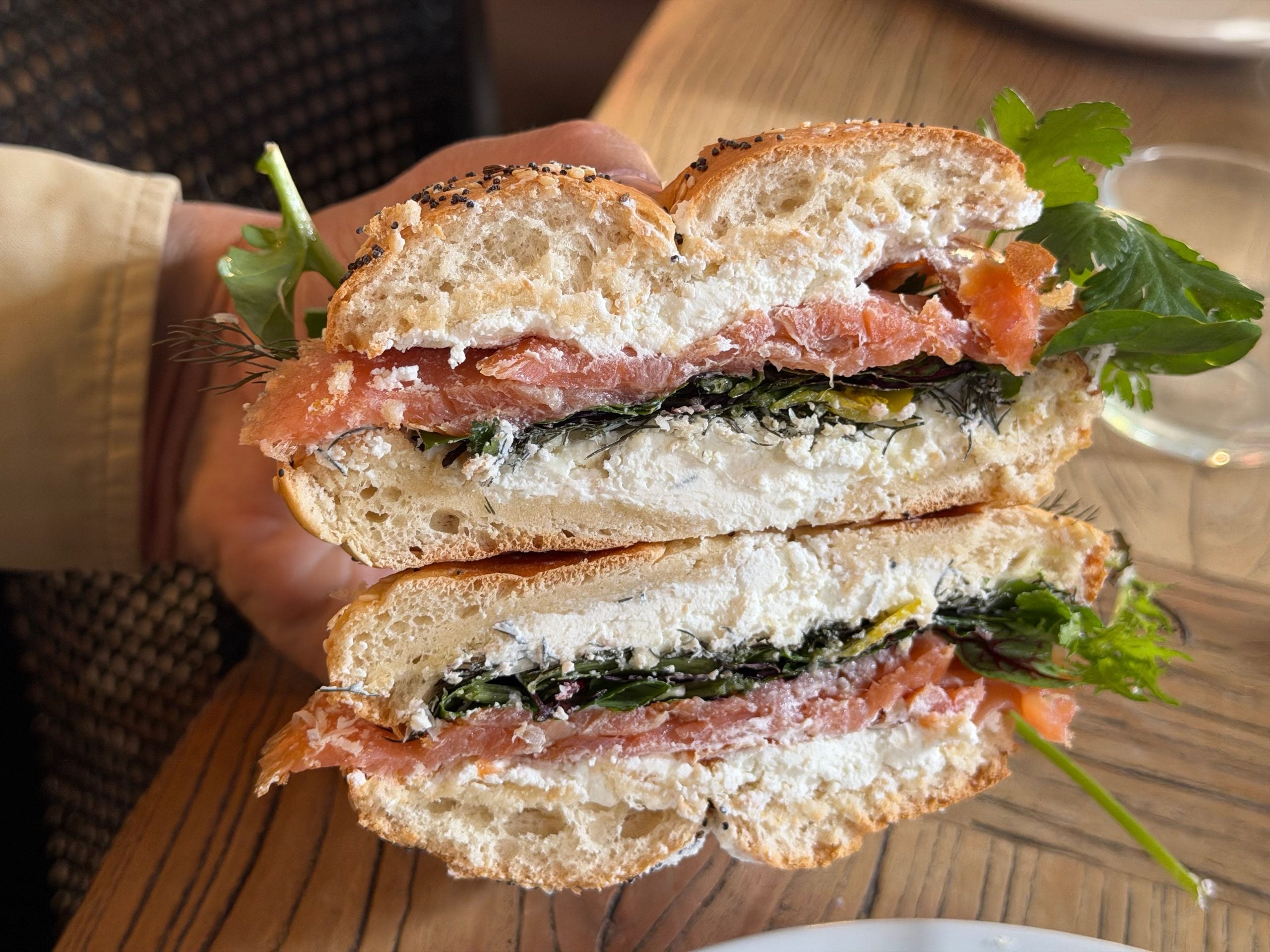 A hand holds a halved bagel sandwich filled with smoked salmon, cream cheese, leafy greens, and herbs, resting on a wooden table.