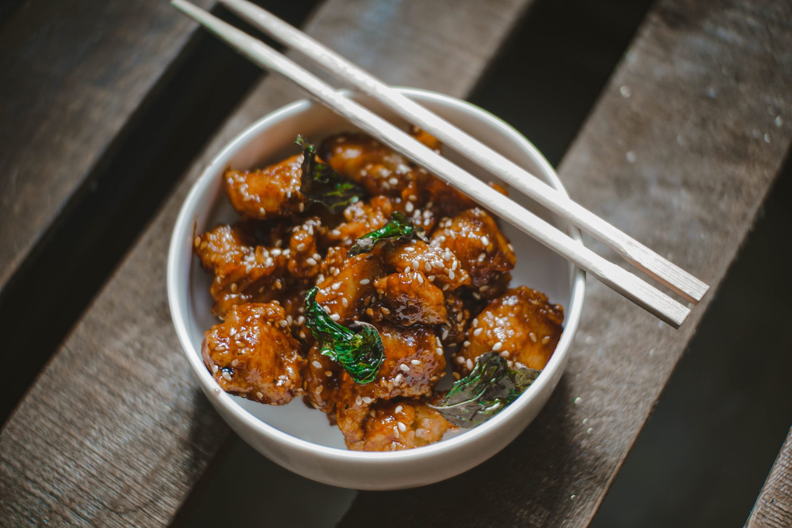 A bowl of glazed, sesame-coated chicken garnished with herbs, served with wooden chopsticks on top, placed on a wooden bench.