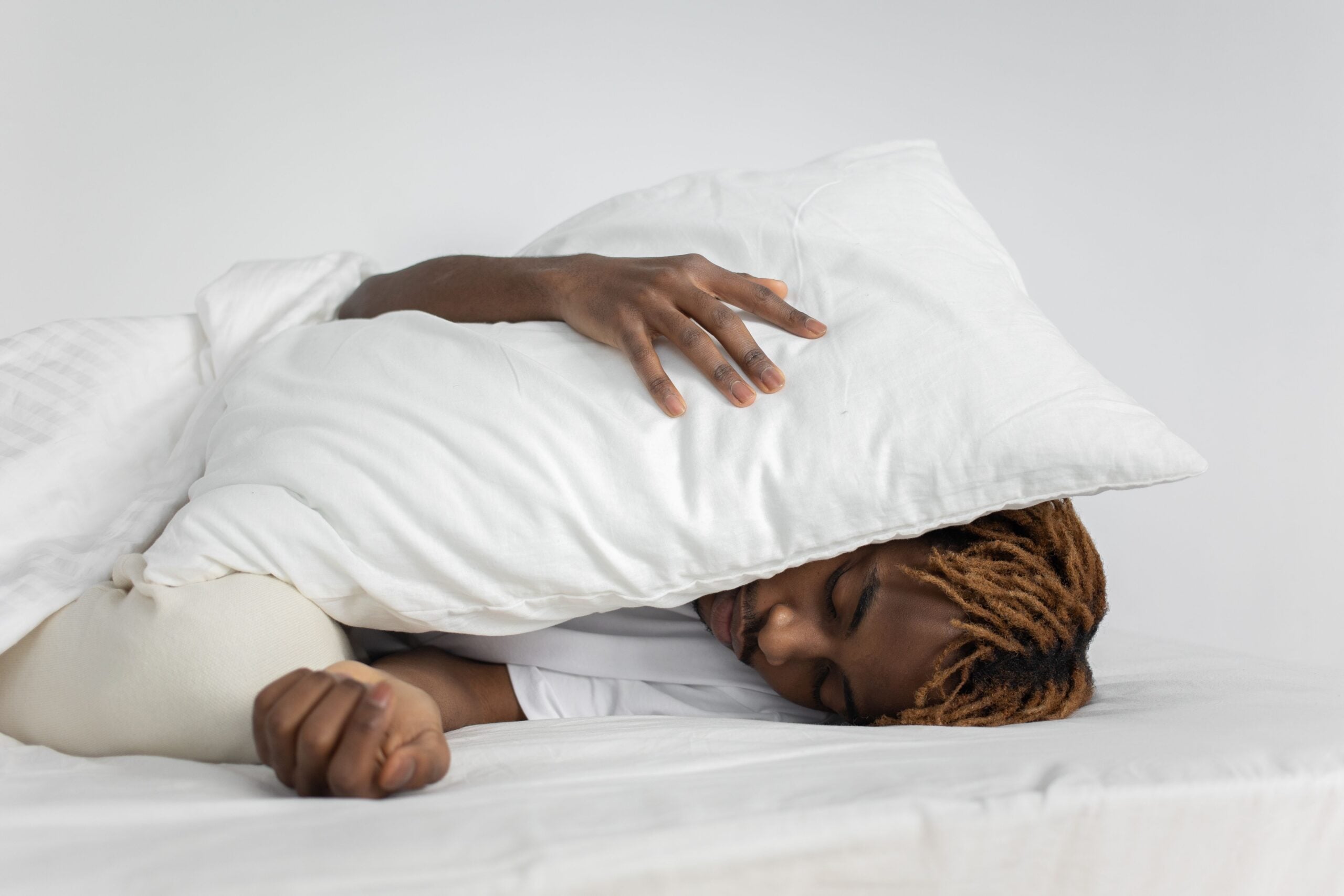 Person lying in bed on their side, hugging a white pillow over their head. They have short, light brown dreadlocks and appear to be resting or sleeping on white bedding against a plain background.