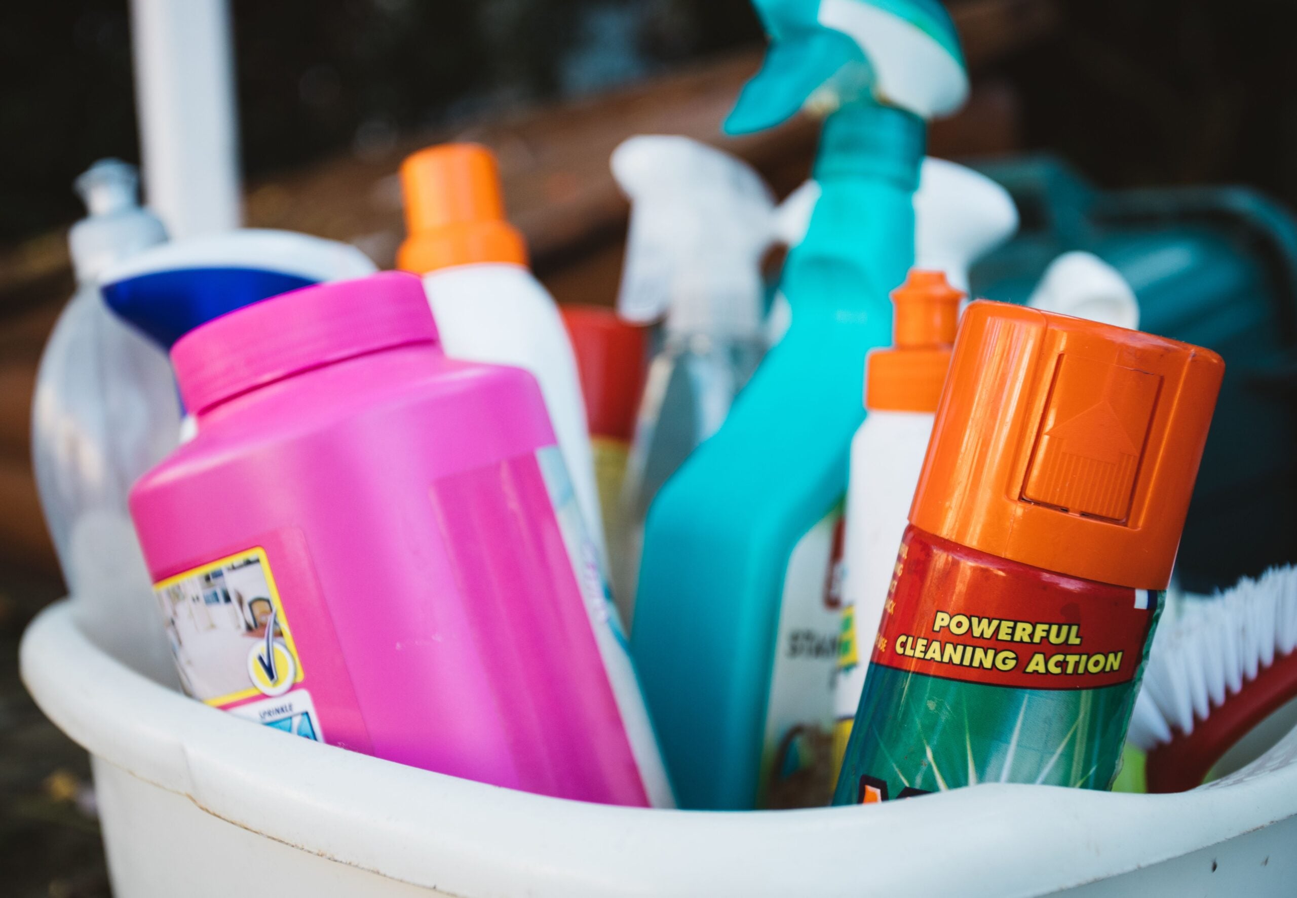 A white bucket filled with various colorful cleaning supplies, including spray bottles, a pink detergent container, and an orange can labeled “Powerful Cleaning Action.”