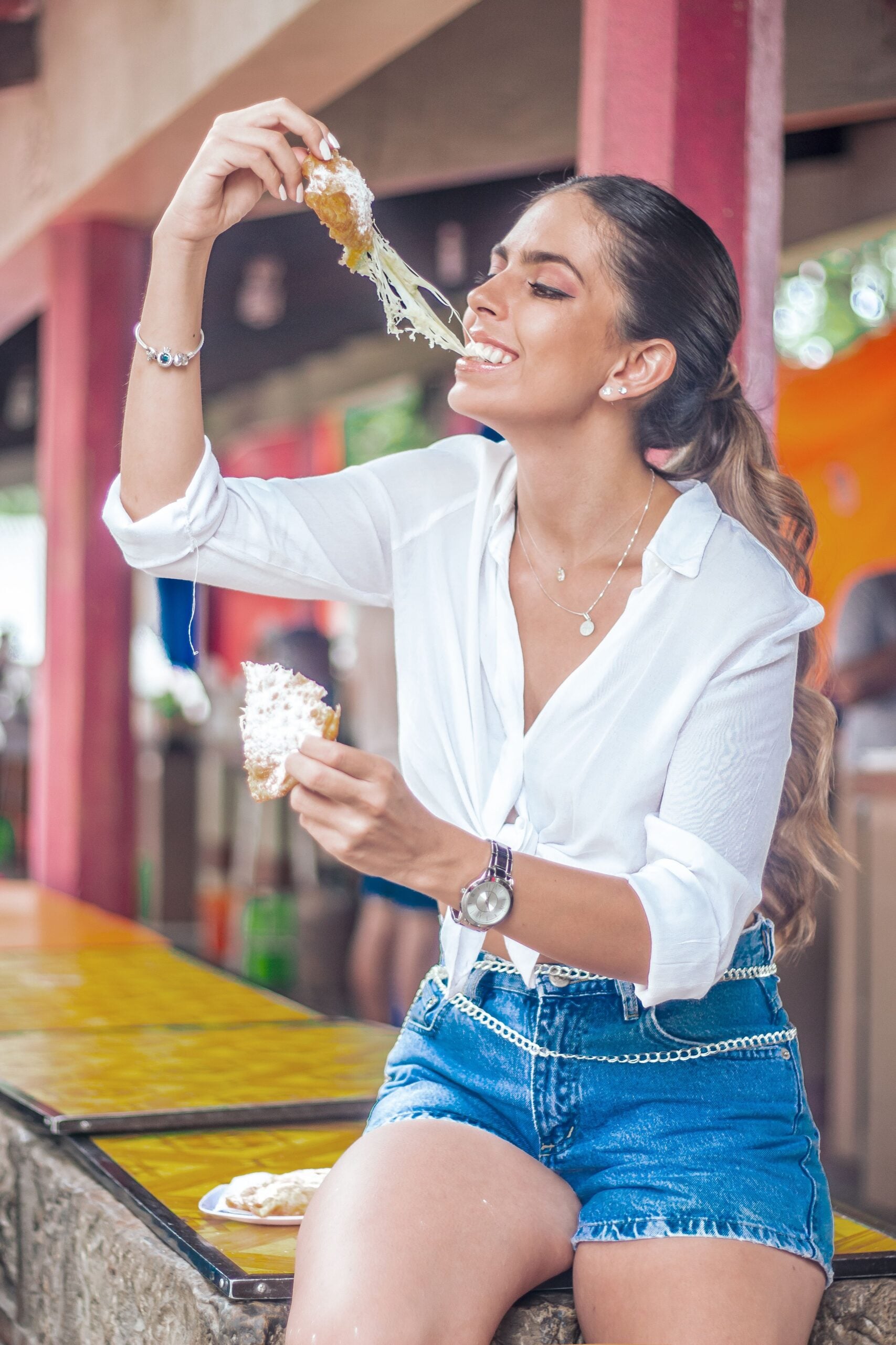 A woman in a white shirt and denim shorts sits, smiling as she lifts a cheesy, saucy food to her mouth, enjoying a meal at what appears to be an outdoor market or food stall.