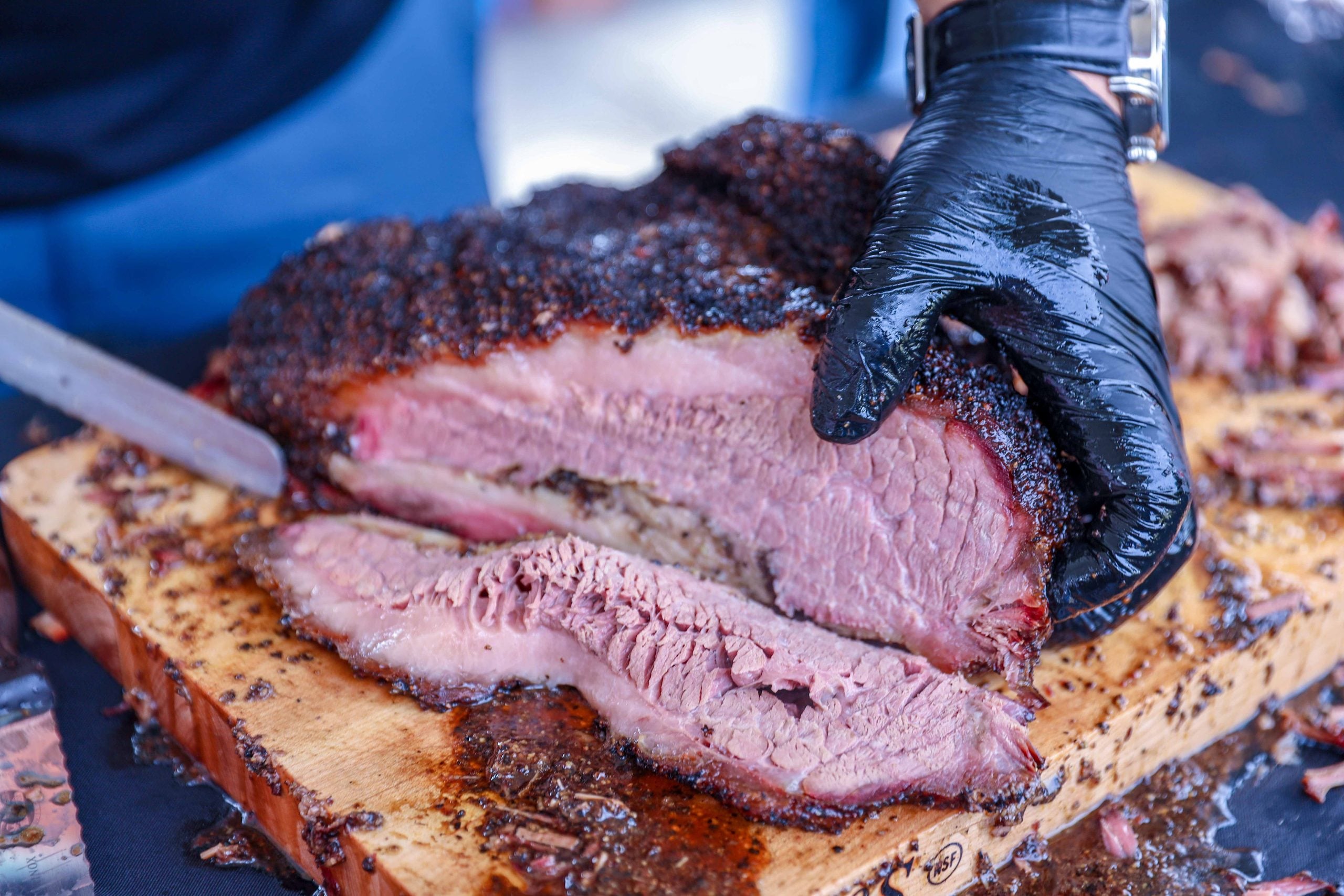 A person wearing a black glove slices a large, juicy smoked brisket—one of the most mouth-watering comfort foods—on a wooden cutting board. The meat has a dark, seasoned crust and tender pink interior.