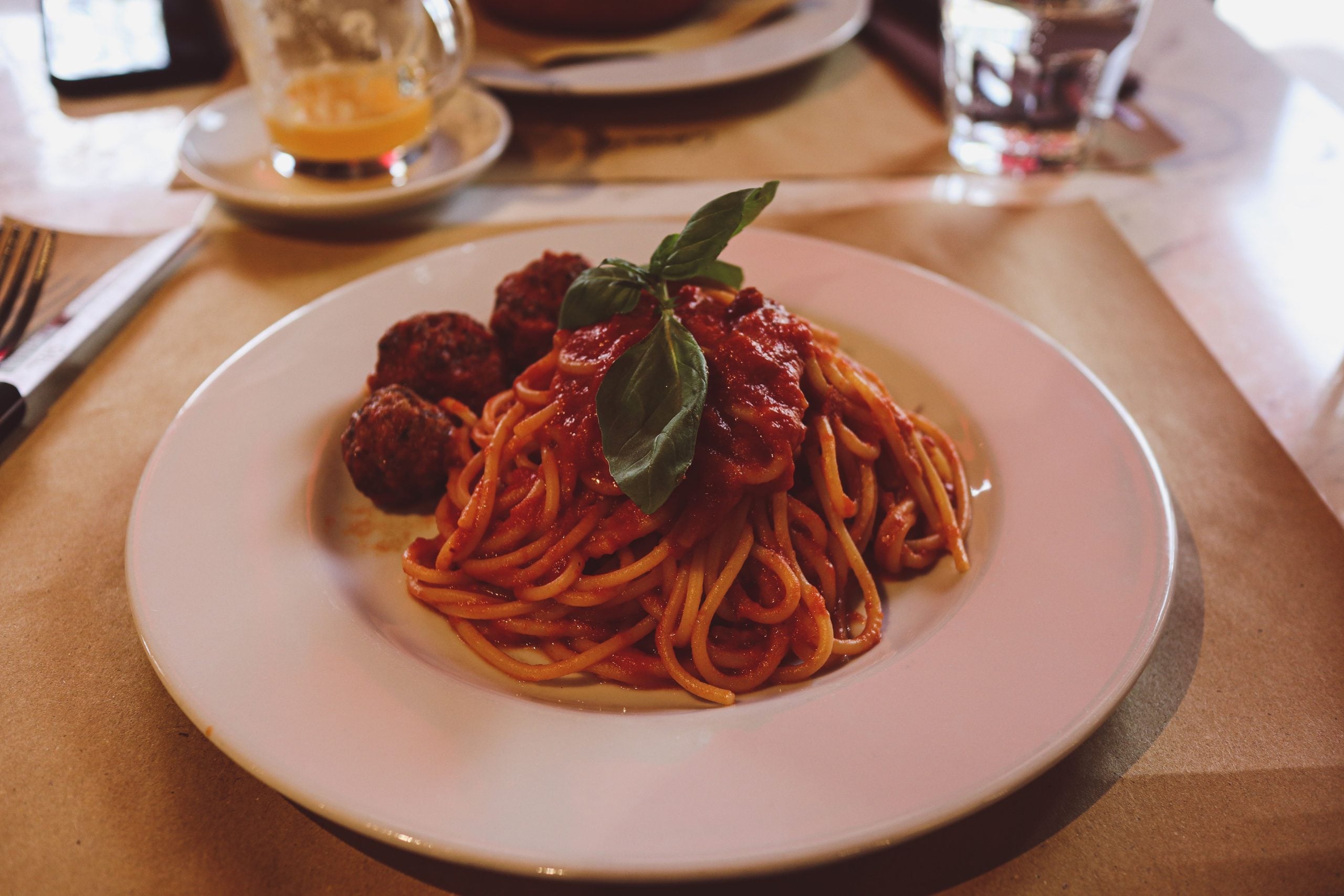 A plate of spaghetti with tomato sauce, garnished with fresh basil and served with three meatballs. A glass and utensils are in the background on a restaurant table.