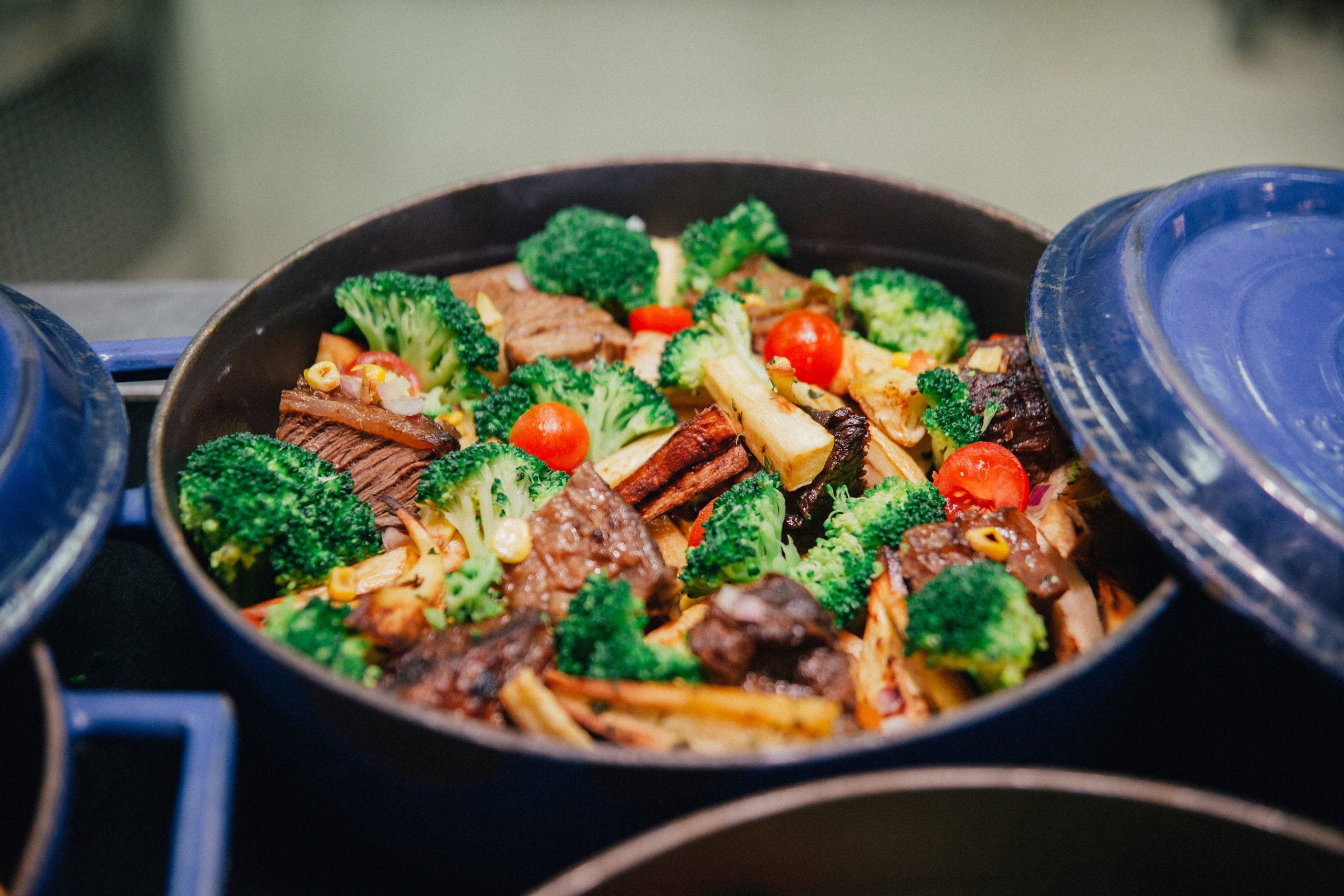 A blue pot filled with cooked broccoli, cherry tomatoes, beef slices, and other vegetables, with its lid partially open.