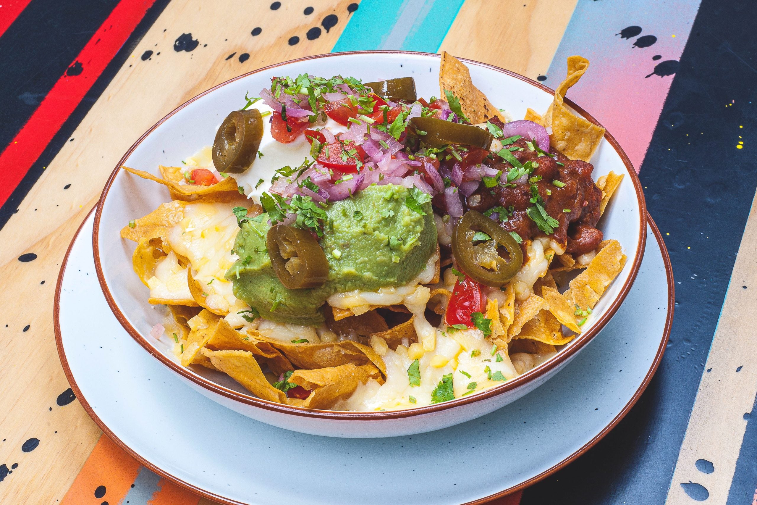 A bowl of nachos topped with melted cheese, guacamole, jalapeños, beans, diced tomatoes, and chopped red onions, served on a colorful, painted table.
