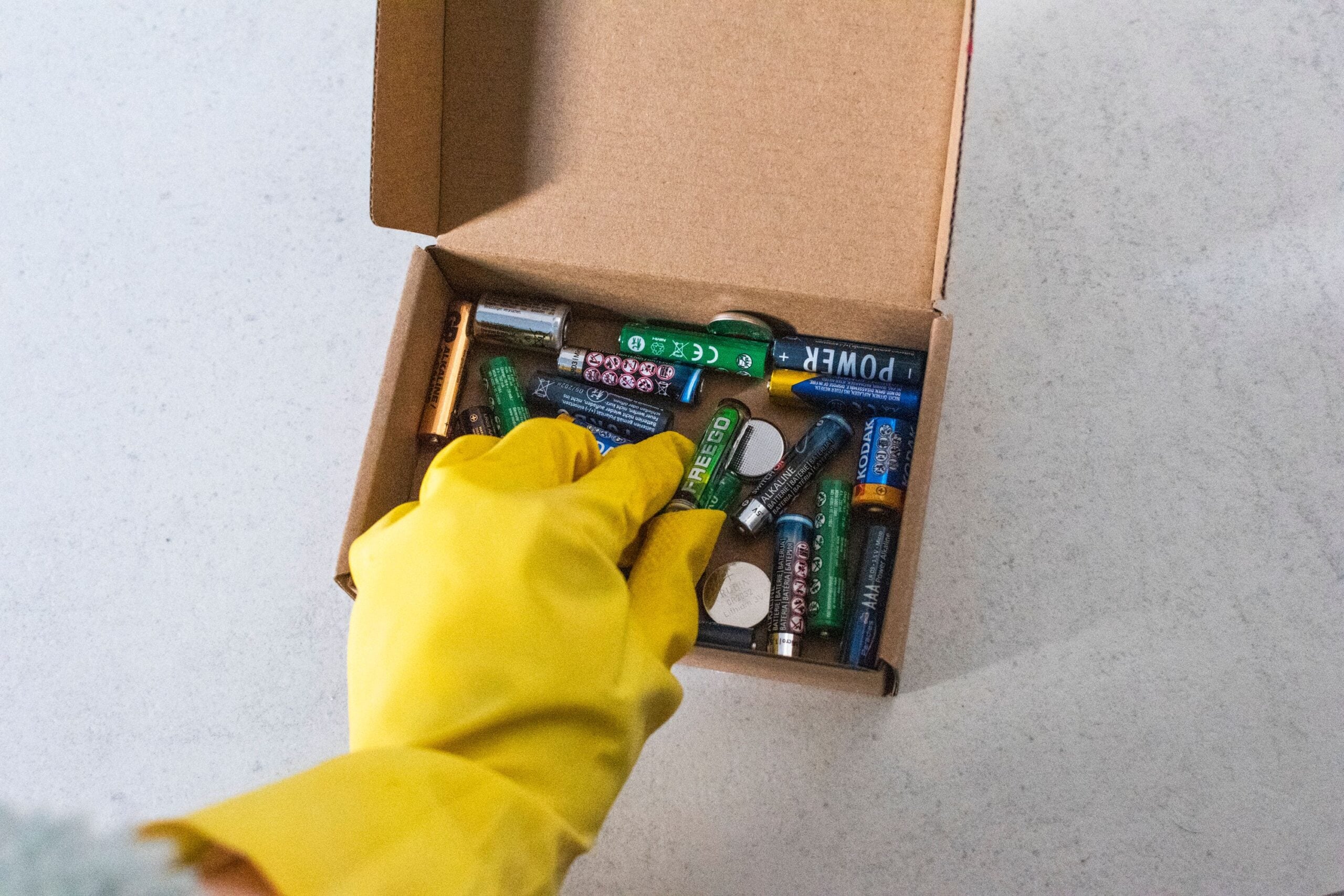 A person wearing a yellow rubber glove picks up a used battery from a cardboard box filled with various old batteries on a light-colored surface.