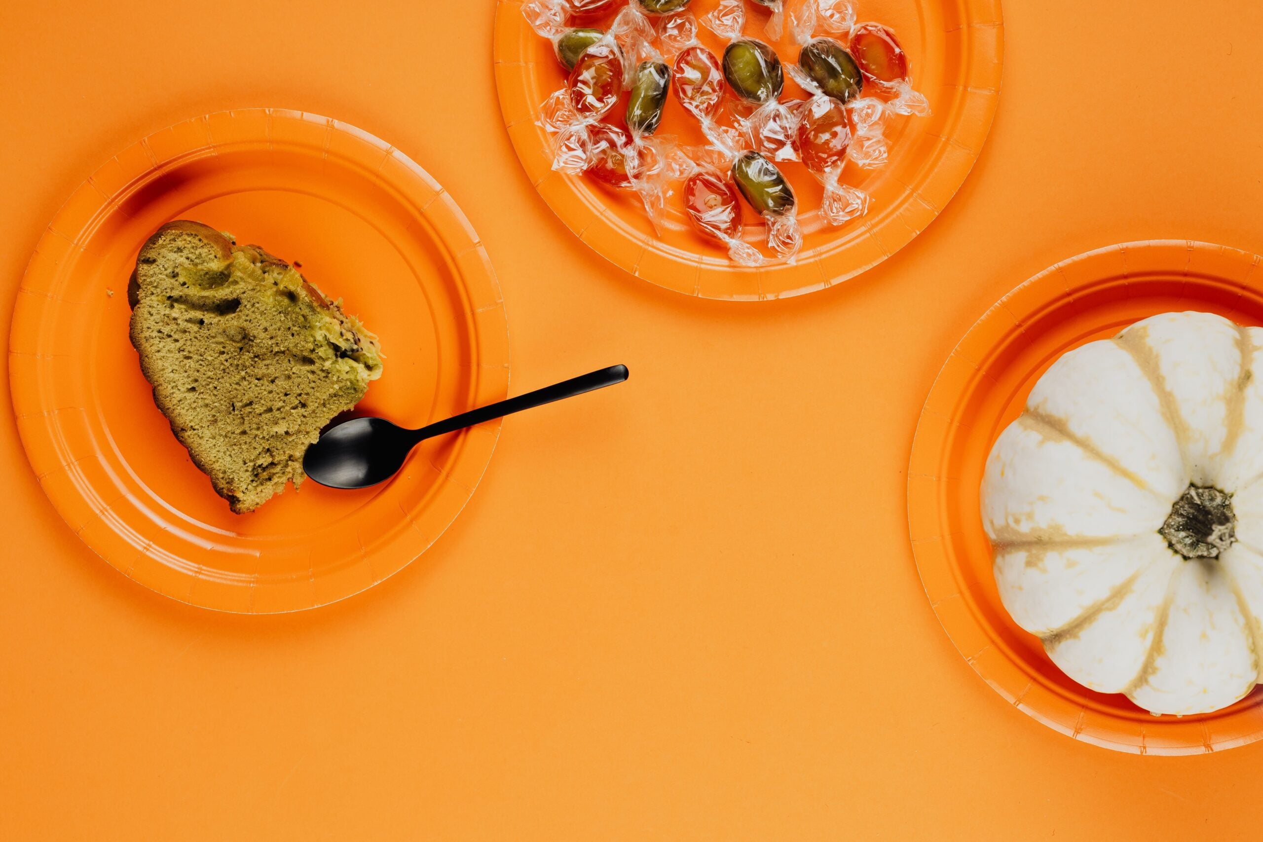 Three orange plates on an orange background: one with a slice of cake and a black spoon, one with wrapped candies, and one with a small white pumpkin.