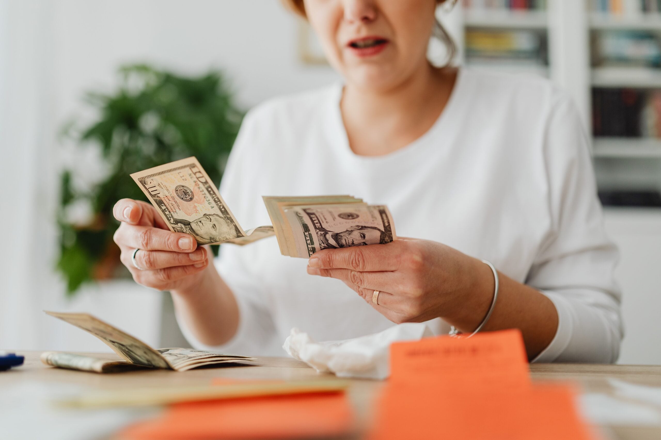 A woman wearing a white shirt is trying to stockpile US dollar bills at a table, with papers and a notebook in front of her, appearing concerned or stressed.