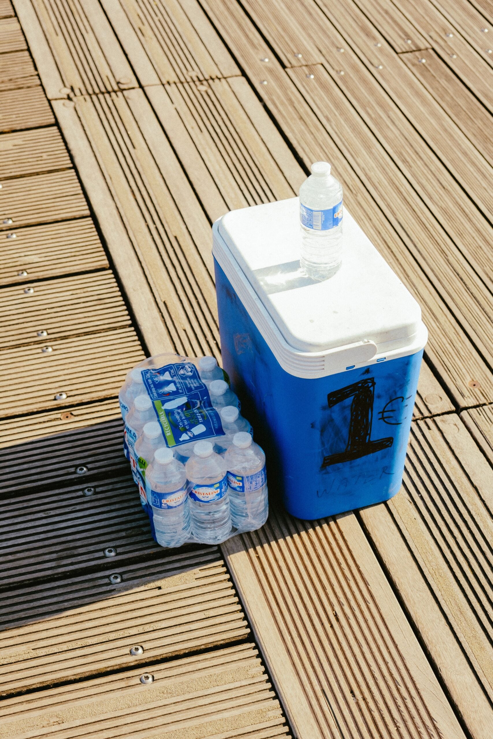 A blue cooler with a large black “1” on it sits on wooden planks, next to a pack of bottled water. A single water bottle is placed on top of the closed cooler.