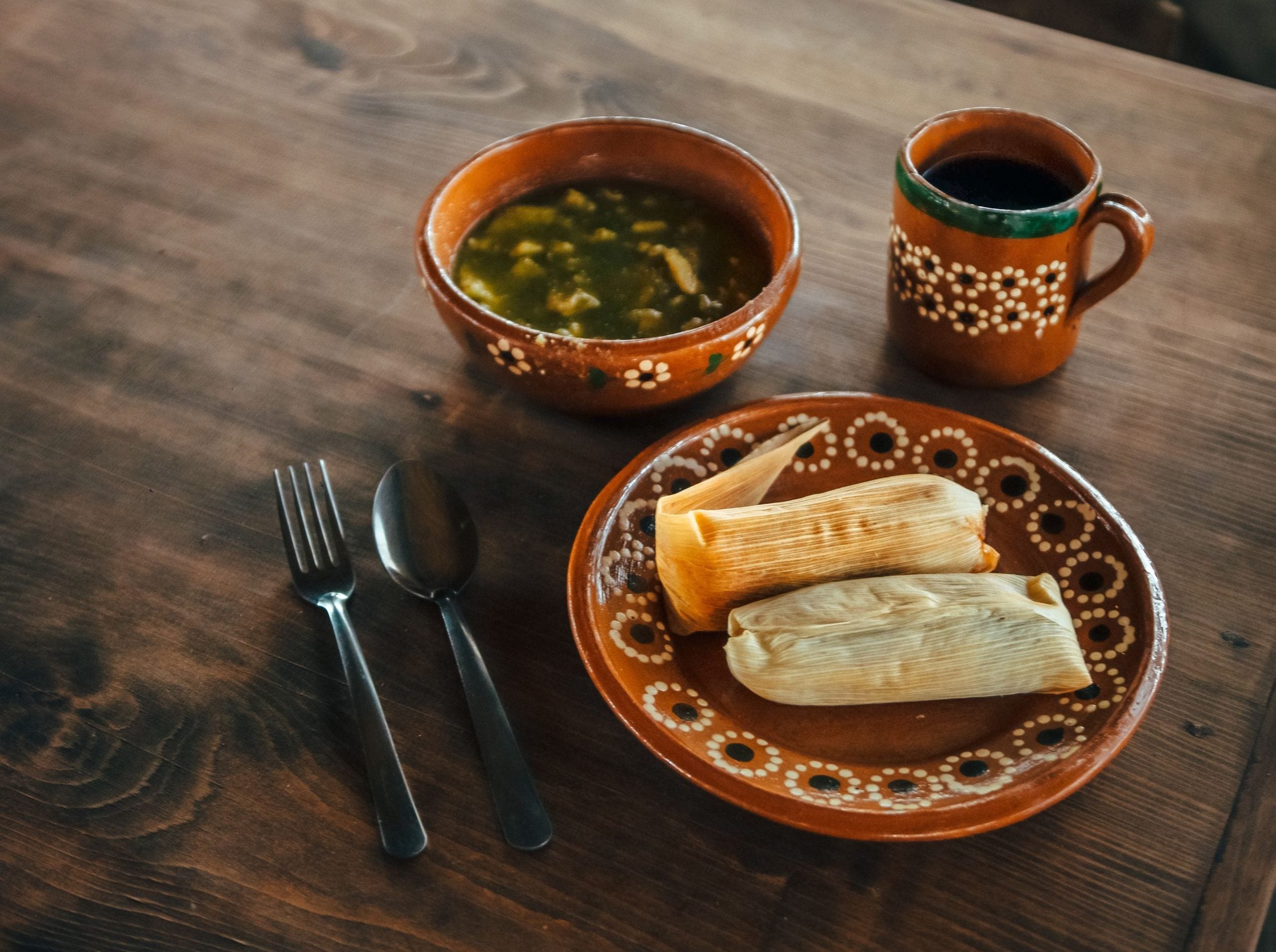 A rustic table set with a plate of two tamales, a bowl of green soup, a mug, and a fork and spoon. These delicious foods are served in traditional brown pottery dishes adorned with white patterns.