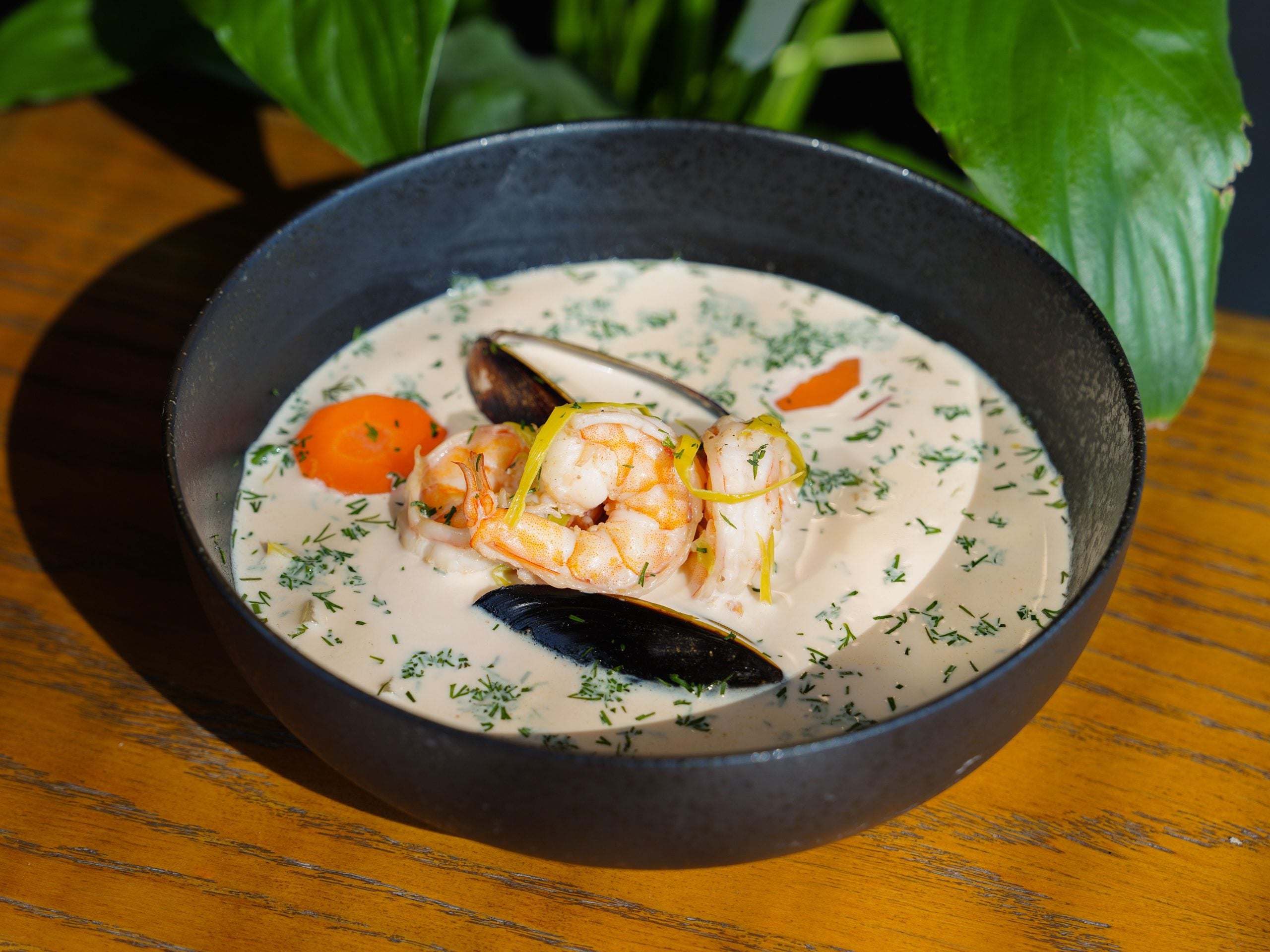 A black bowl of creamy seafood soup, one of the most comforting foods, with shrimp, mussels, carrot slices, and herbs sits on a wooden table with green leaves in the background.