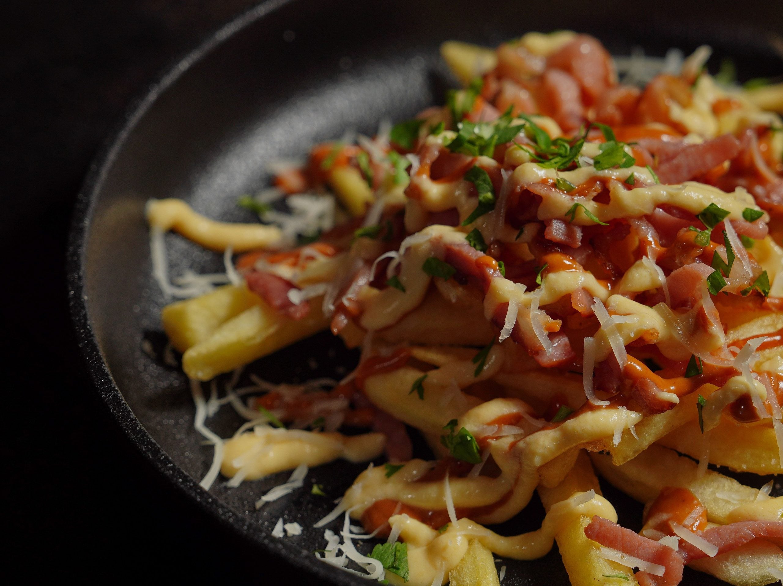 A close-up of loaded fries topped with melted cheese, chopped ham, sauce, and sprinkled herbs, served in a black dish—an irresistible combination that showcases the best of comfort foods.