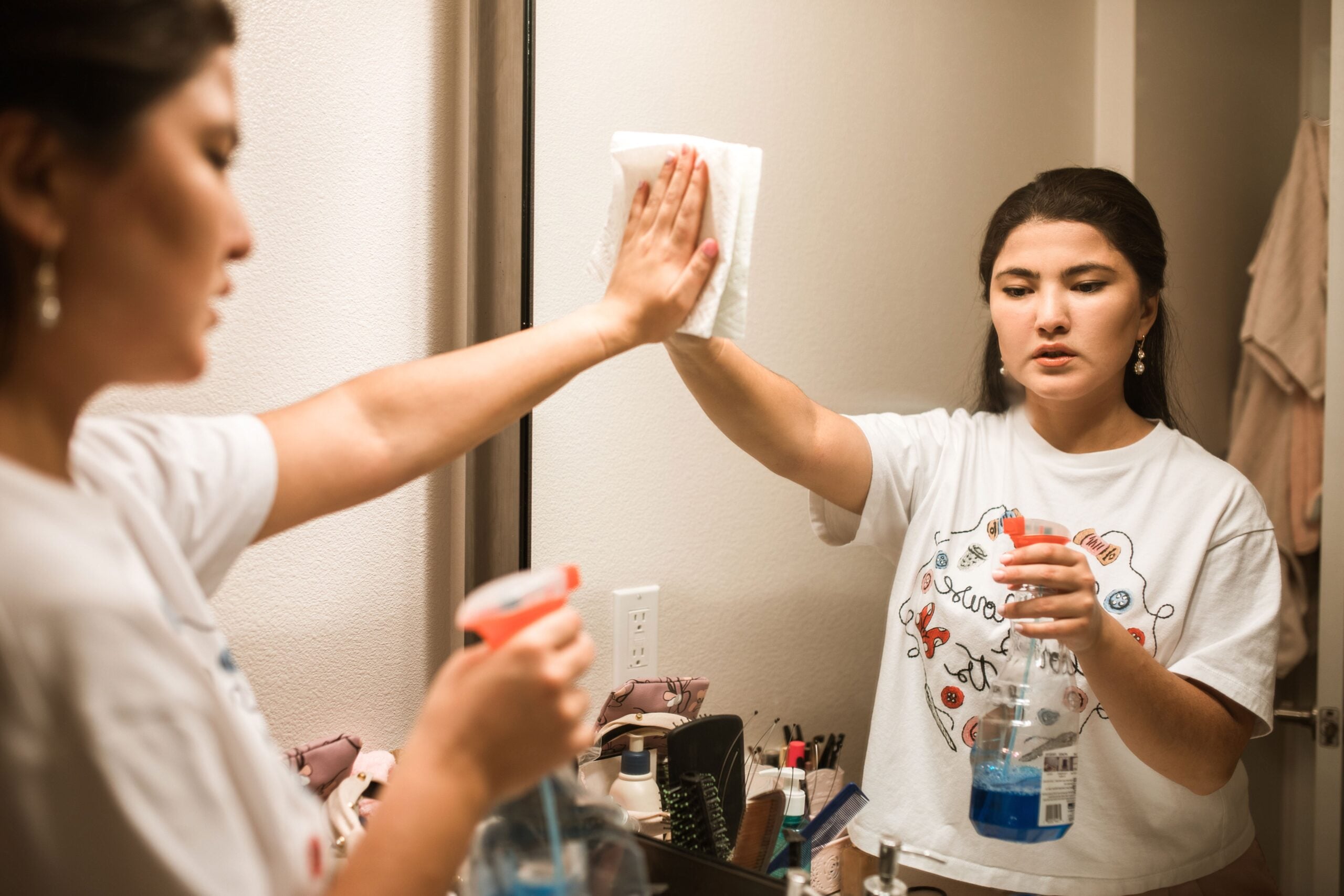 A woman in a white t-shirt cleans a bathroom mirror using a spray bottle and a cloth. She stands at the sink, focusing on wiping the glass, with various toiletries visible on the counter.