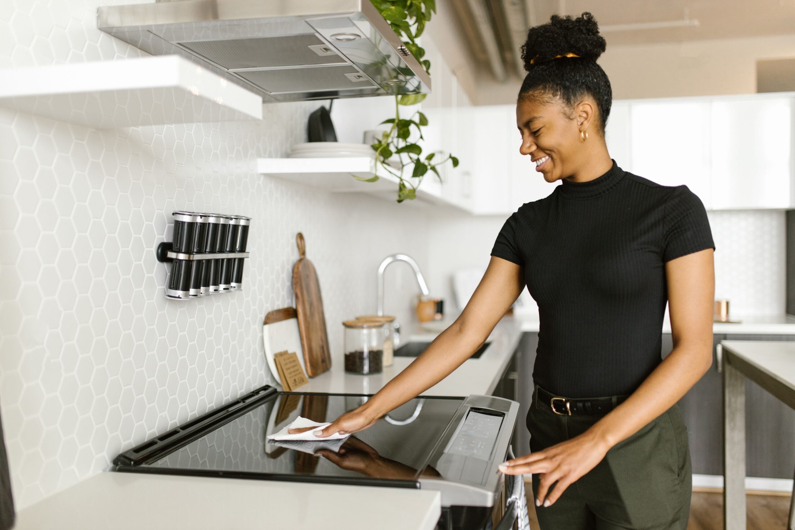 A woman in a black shirt smiles while cleaning a stovetop in a modern kitchen with white hexagon tiles and wooden accents.