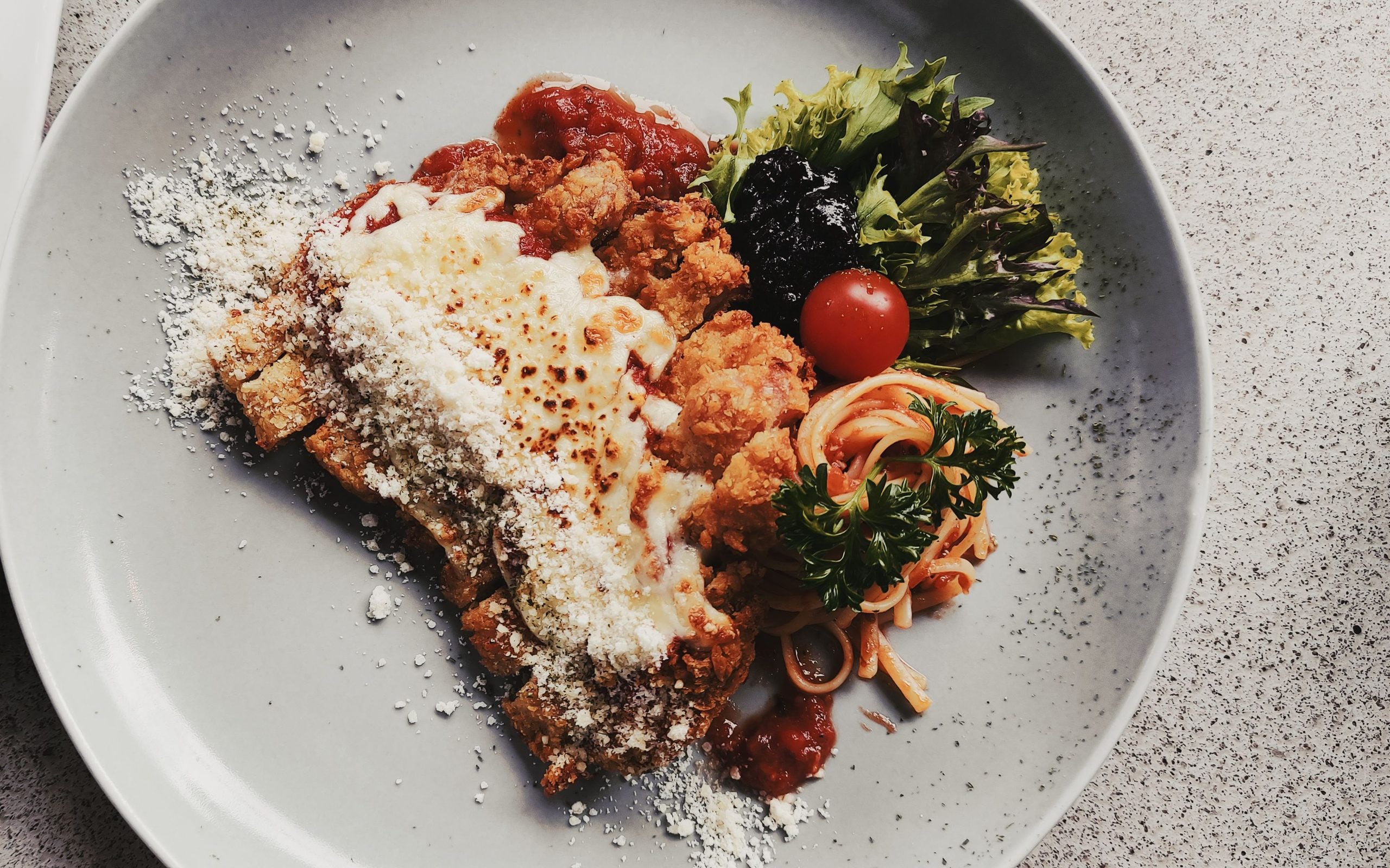 A plate with breaded chicken topped with cheese and tomato sauce, served with spaghetti, a side salad with lettuce and a cherry tomato, and garnished with grated cheese.