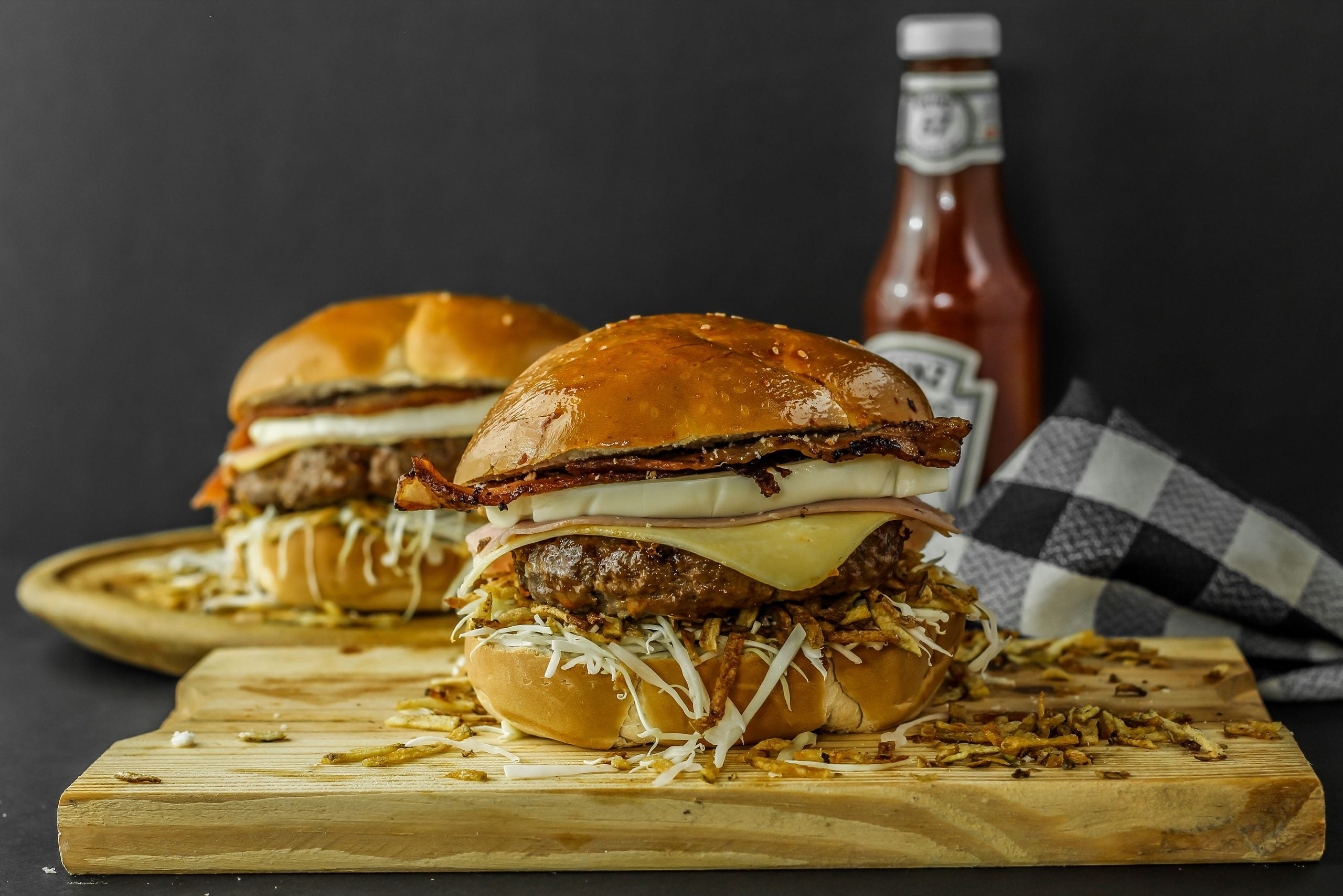 Two cheeseburgers with lettuce, cheese, and bacon—classic comfort foods—sit on a wooden board. A bottle of ketchup and a black-and-white checkered napkin are in the background.