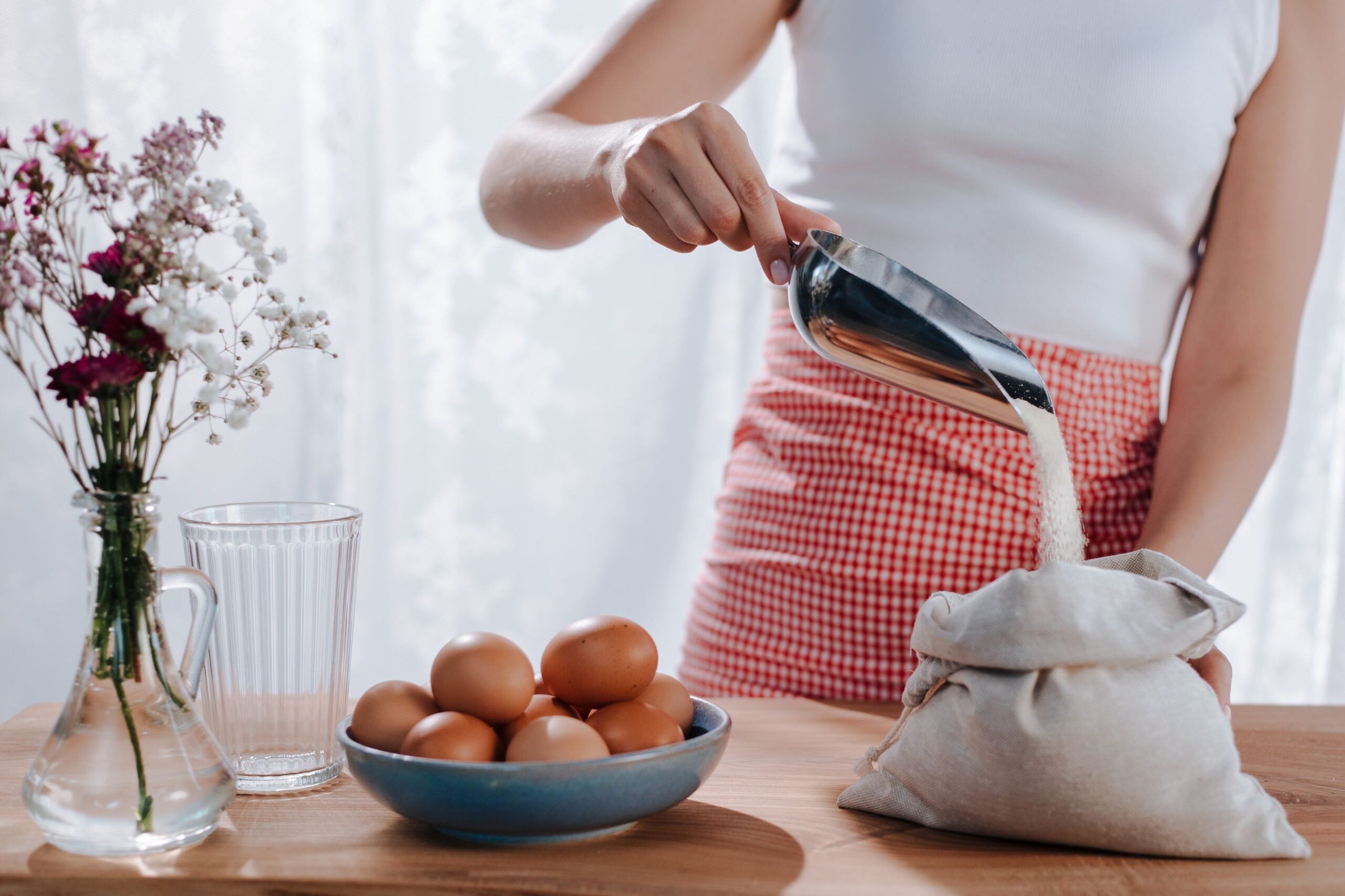 A person in a white top and red checkered apron scoops sugar from a sack on a wooden table with a glass of water, a bowl of eggs, and a vase of flowers.