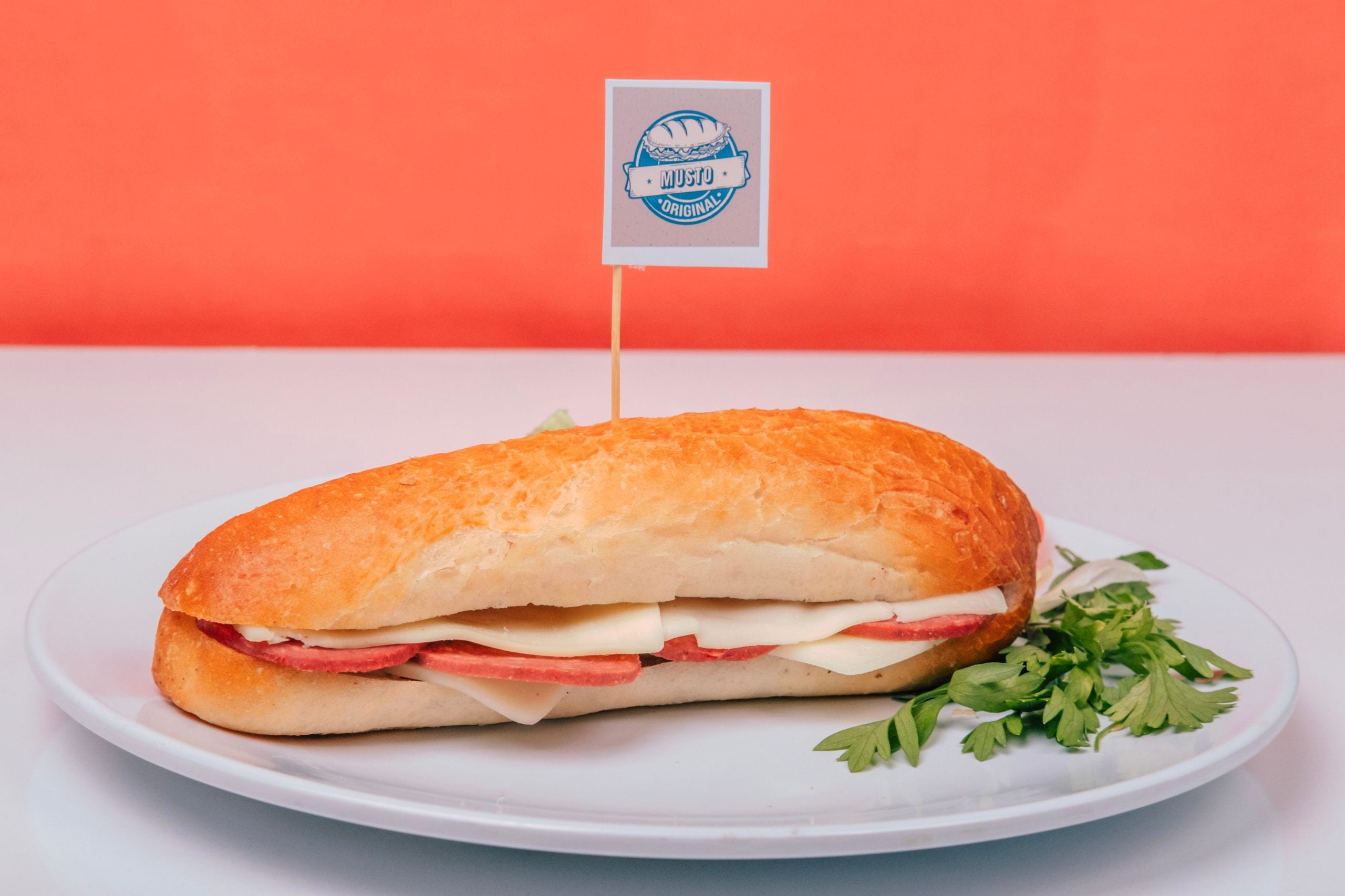 A sandwich with meat and cheese, classic comfort foods, rests on a white plate garnished with parsley in front of an orange background. A small flag with a logo is stuck into the sandwich.