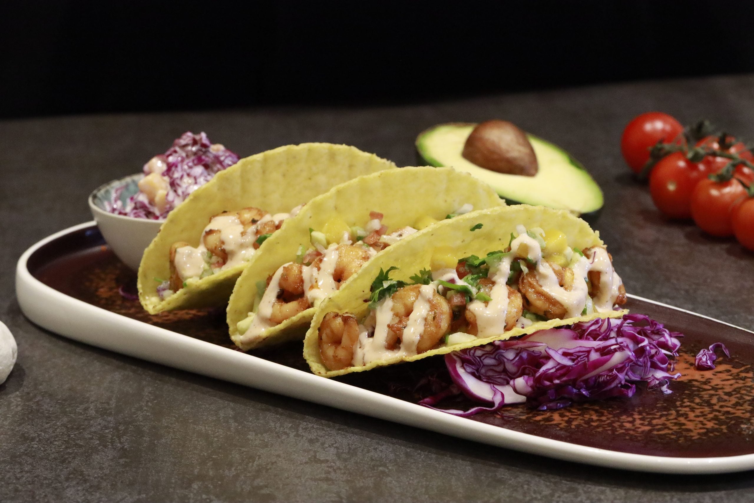 Three tacos filled with grilled chicken, diced onions, cilantro, and a creamy sauce are served on a plate with shredded purple cabbage, with a bowl of slaw, half an avocado, and cherry tomatoes in the background.