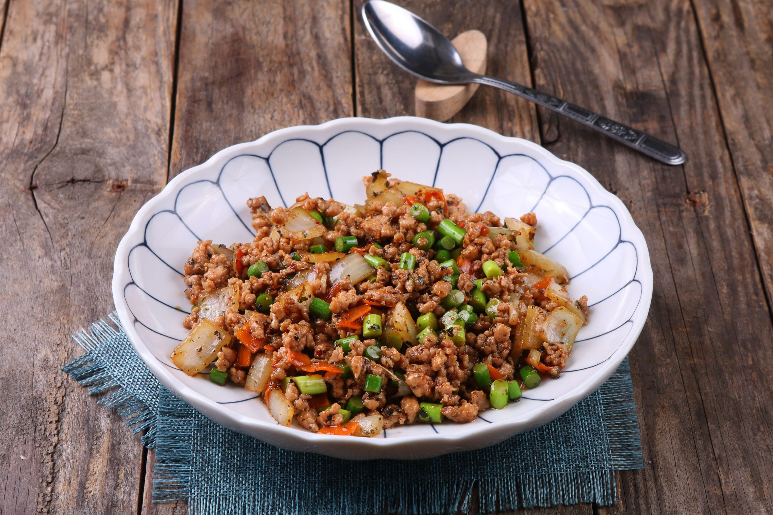 A bowl of stir-fried minced meat with chopped green onions, carrots, and vegetables sits on a blue cloth on a rustic wooden table, with a spoon in the background.