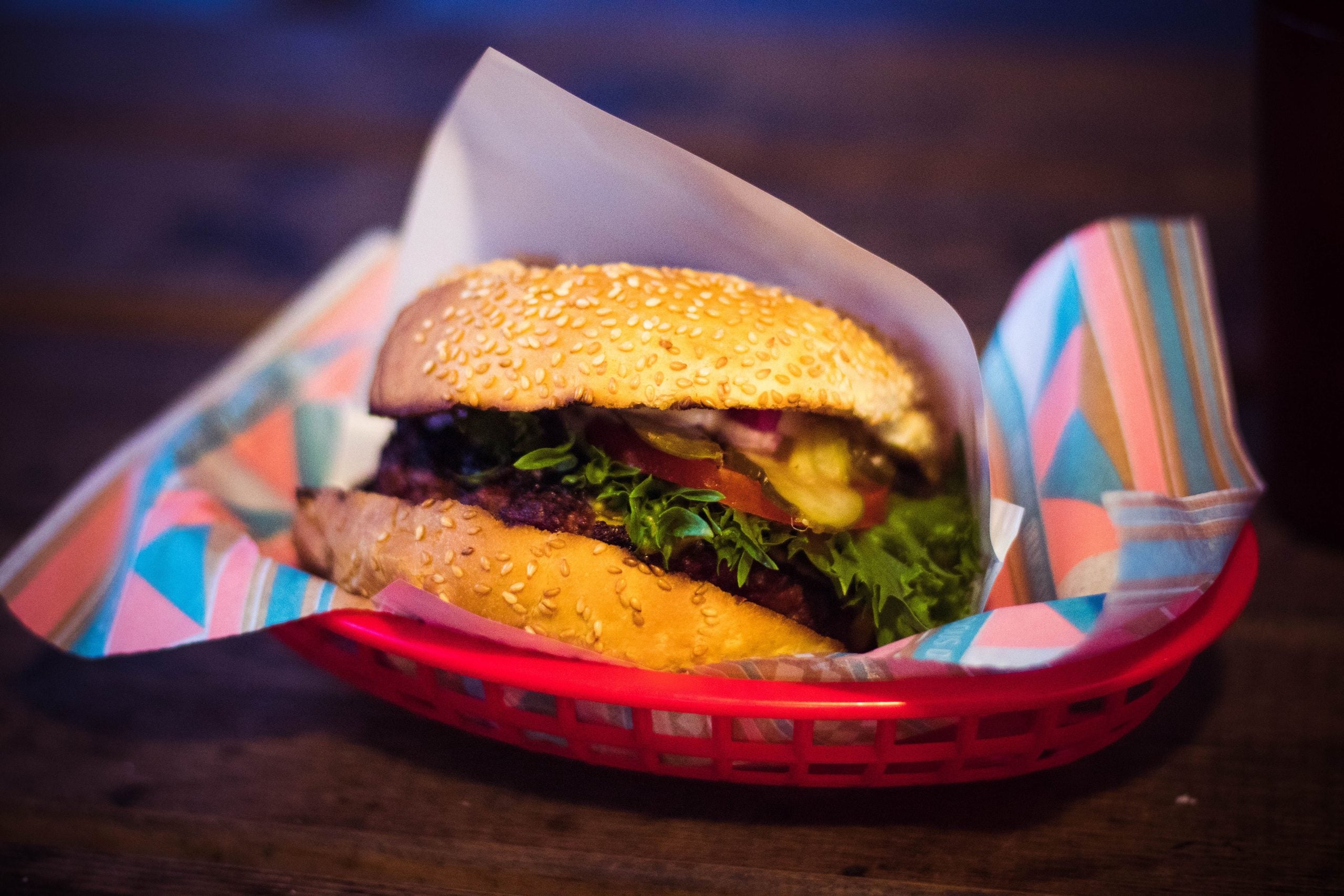 A sesame seed hamburger with lettuce, tomato, and onions sits in a red plastic basket lined with colorful paper on a wooden table.