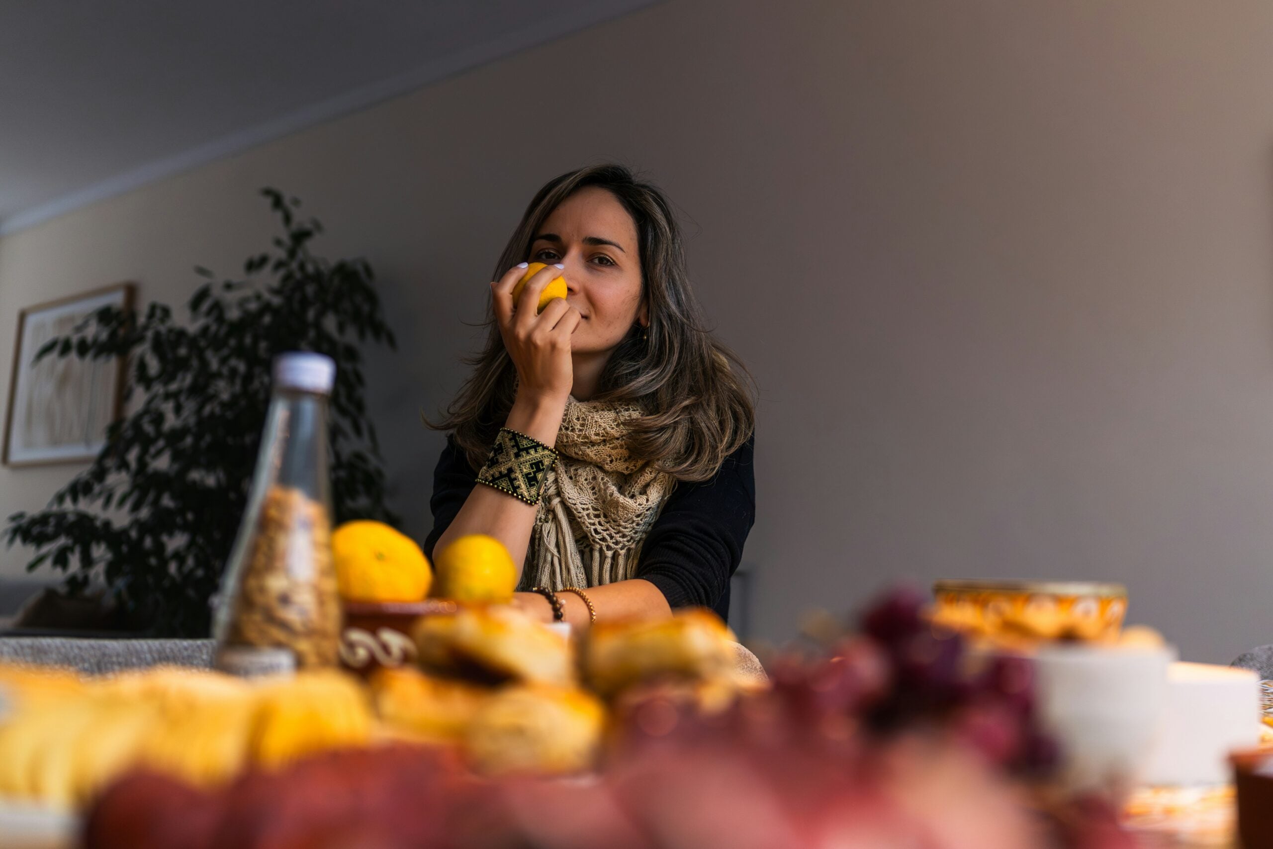 A woman sits at a table with assorted food, holding a yellow fruit near her face, smiling softly. The foreground shows blurred fruit and a bottle, while a plant is visible in the background.