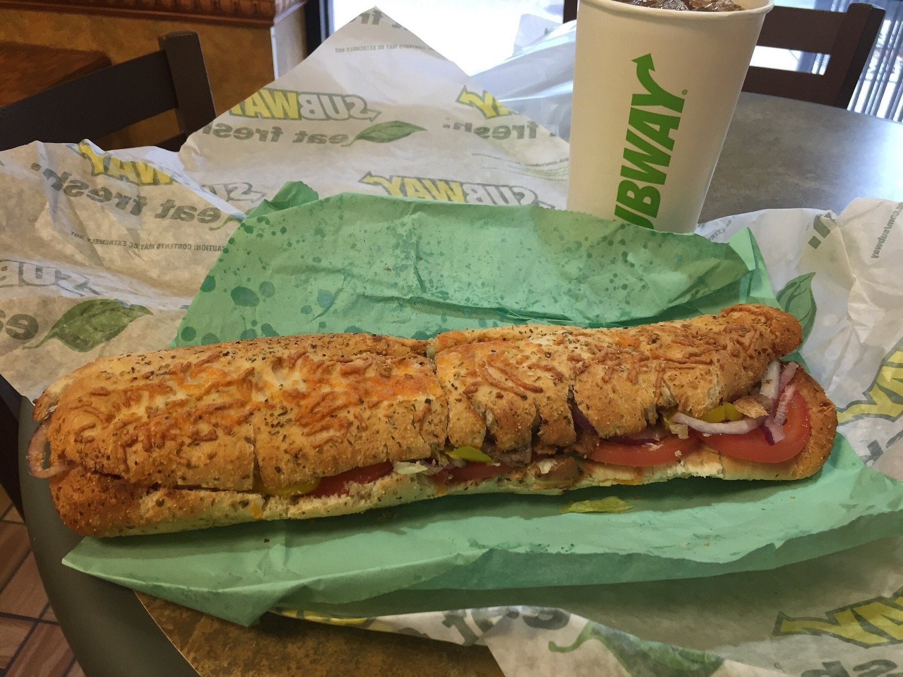A footlong Subway sandwich with vegetables and cheese on a sub roll sits on green sandwich paper, next to a Subway cup filled with a dark soda on a table.