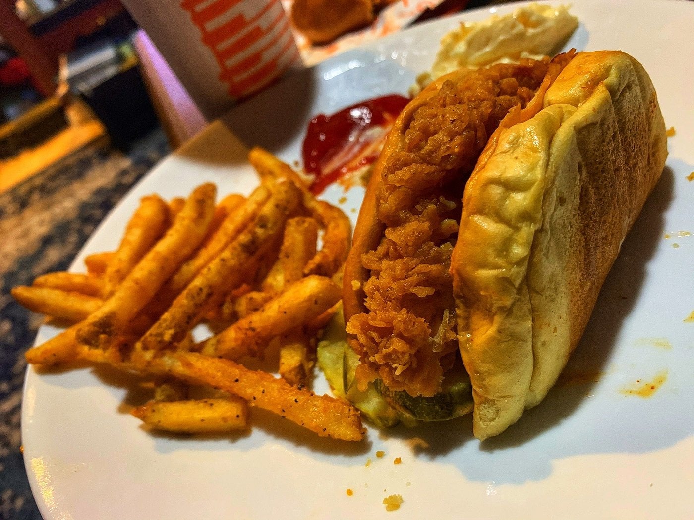 One of the fast-food meals with seasoned fries, a fried chicken sandwich on a bun with pickles, and some ketchup on the side. A Whataburger cup and coleslaw are visible in the background.