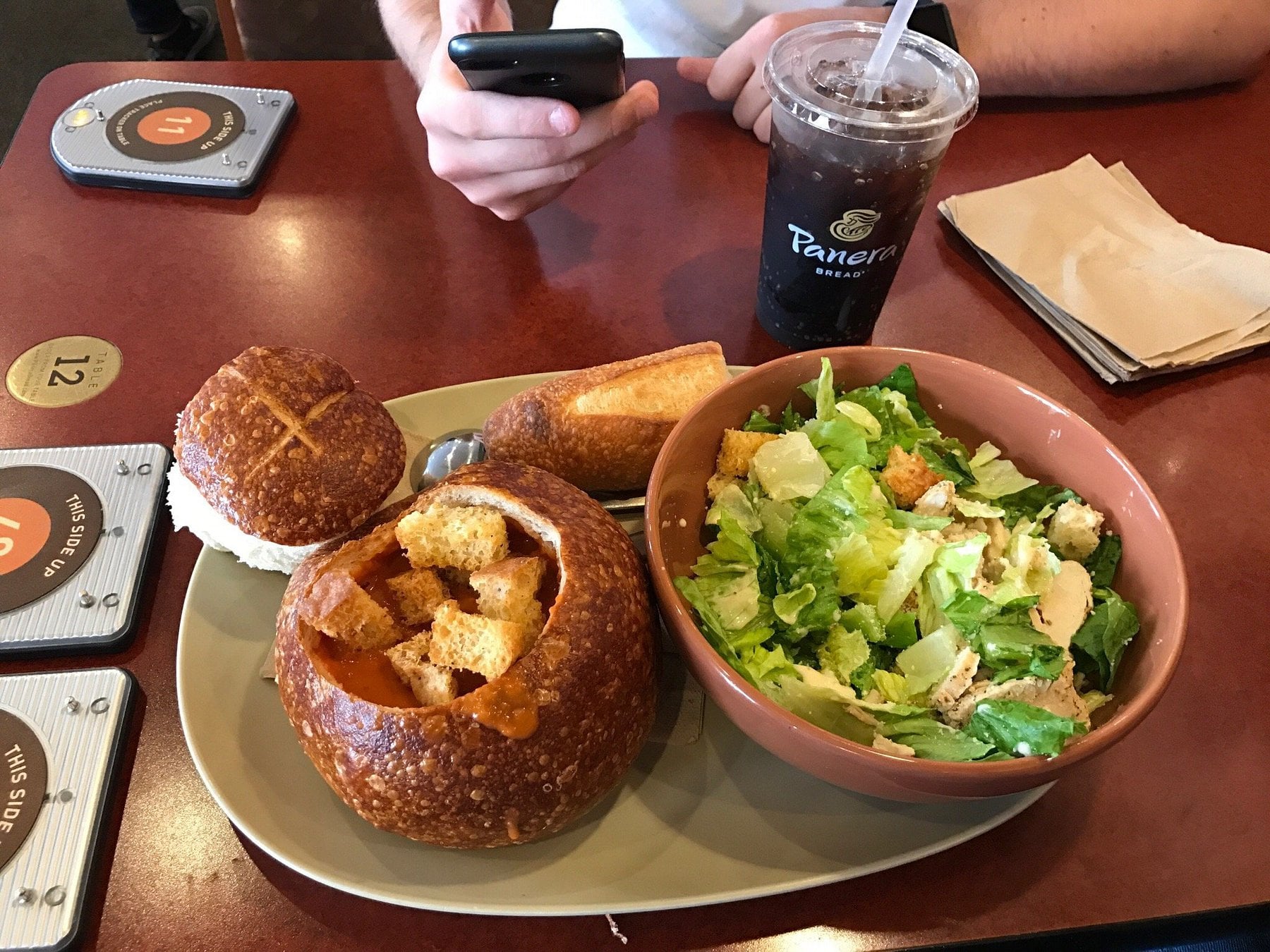 A meal at Panera Bread with a bread bowl of soup, a Caesar salad, a baguette slice, and a drink. A person is holding a phone at the table.