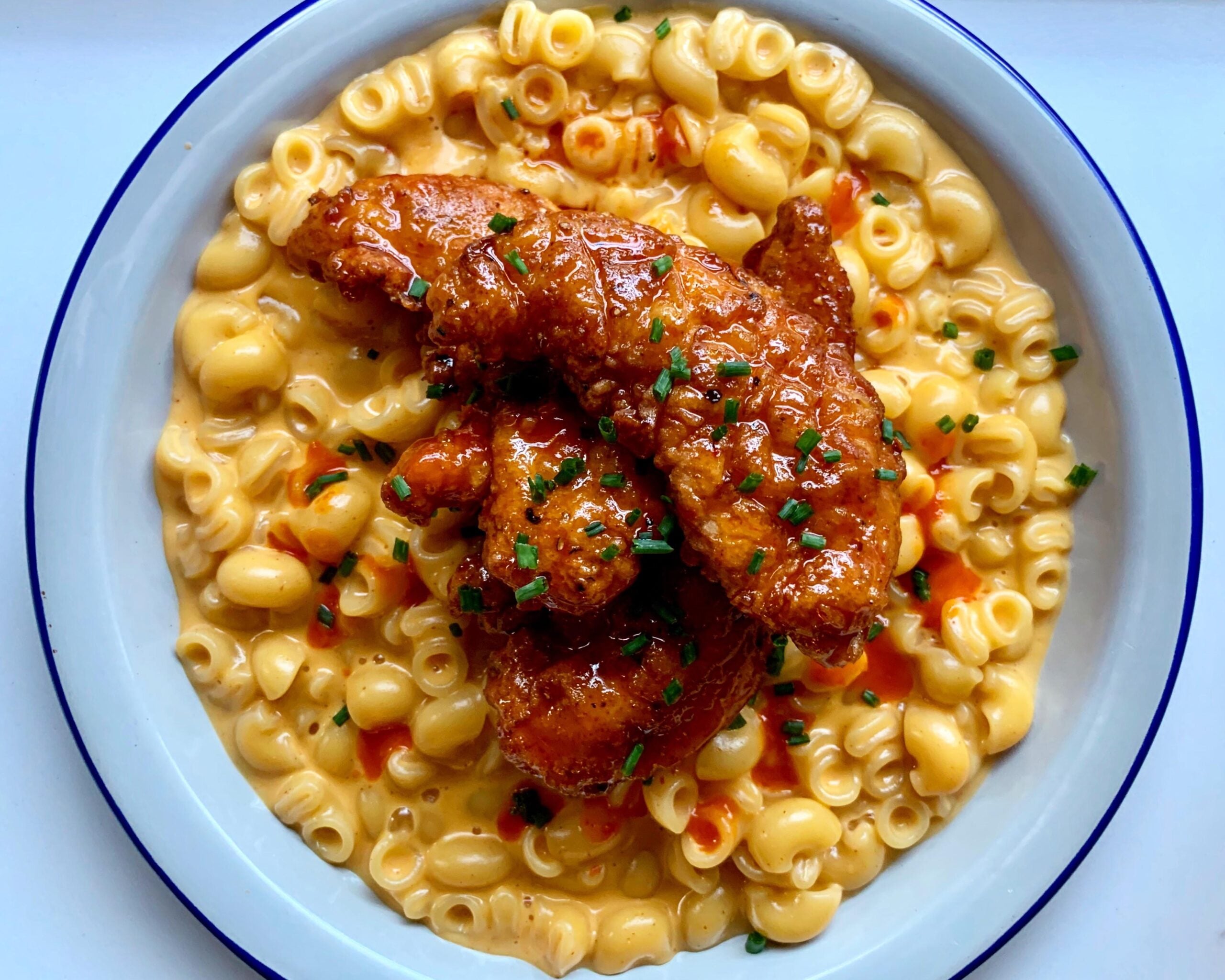 A bowl of creamy macaroni and cheese topped with three pieces of saucy fried chicken tenders, garnished with chopped chives and a drizzle of hot sauce.