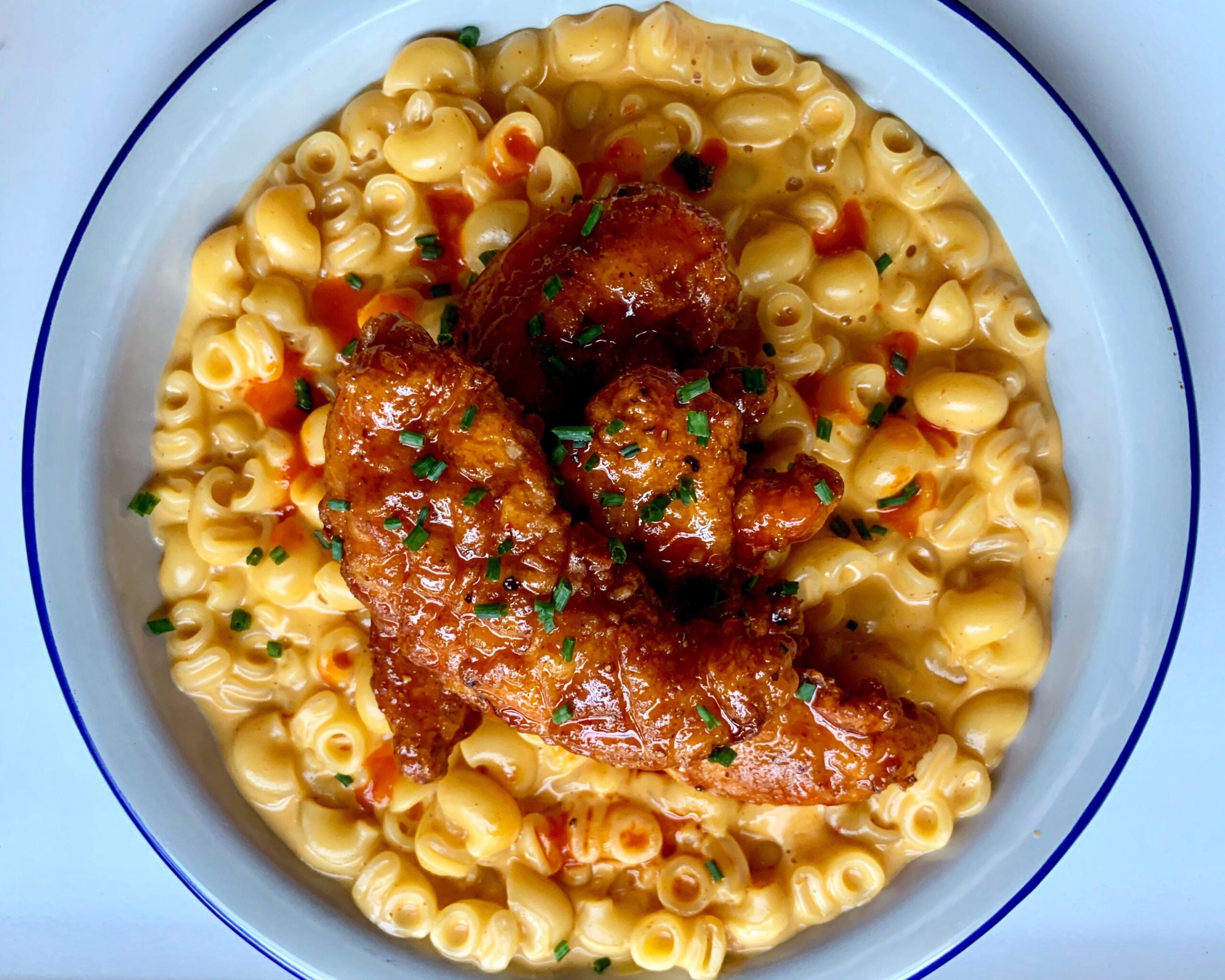 A plate of creamy macaroni and cheese topped with crispy fried chicken tenders, drizzled with hot sauce and sprinkled with chopped herbs.