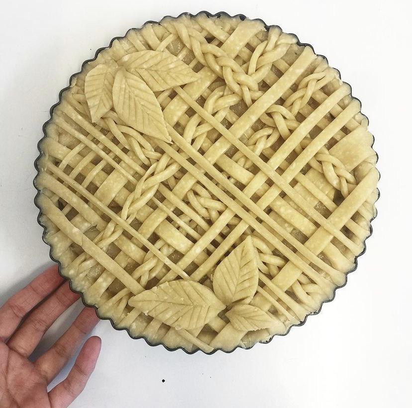 A hand holds a pie with an intricate unbaked lattice crust, featuring braided dough strips and decorative leaf shapes on top, against a white background.