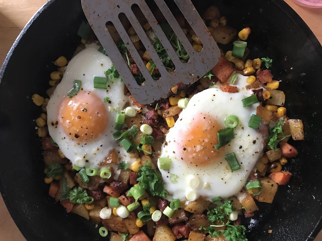 Two sunny-side-up eggs on a skillet hash with potatoes, corn, chopped green onions, parsley, and sausage pieces. A spatula rests above one egg.