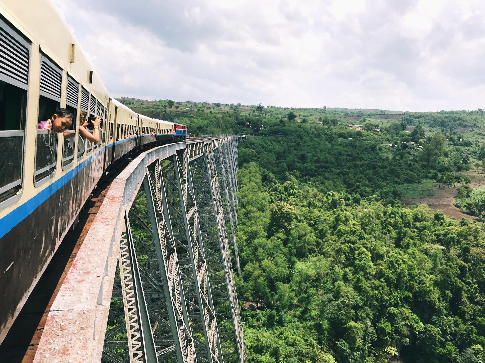 A train crosses a tall, curved steel bridge over a lush green valley, with passengers leaning out the windows to enjoy the scenic view under a partly cloudy sky.