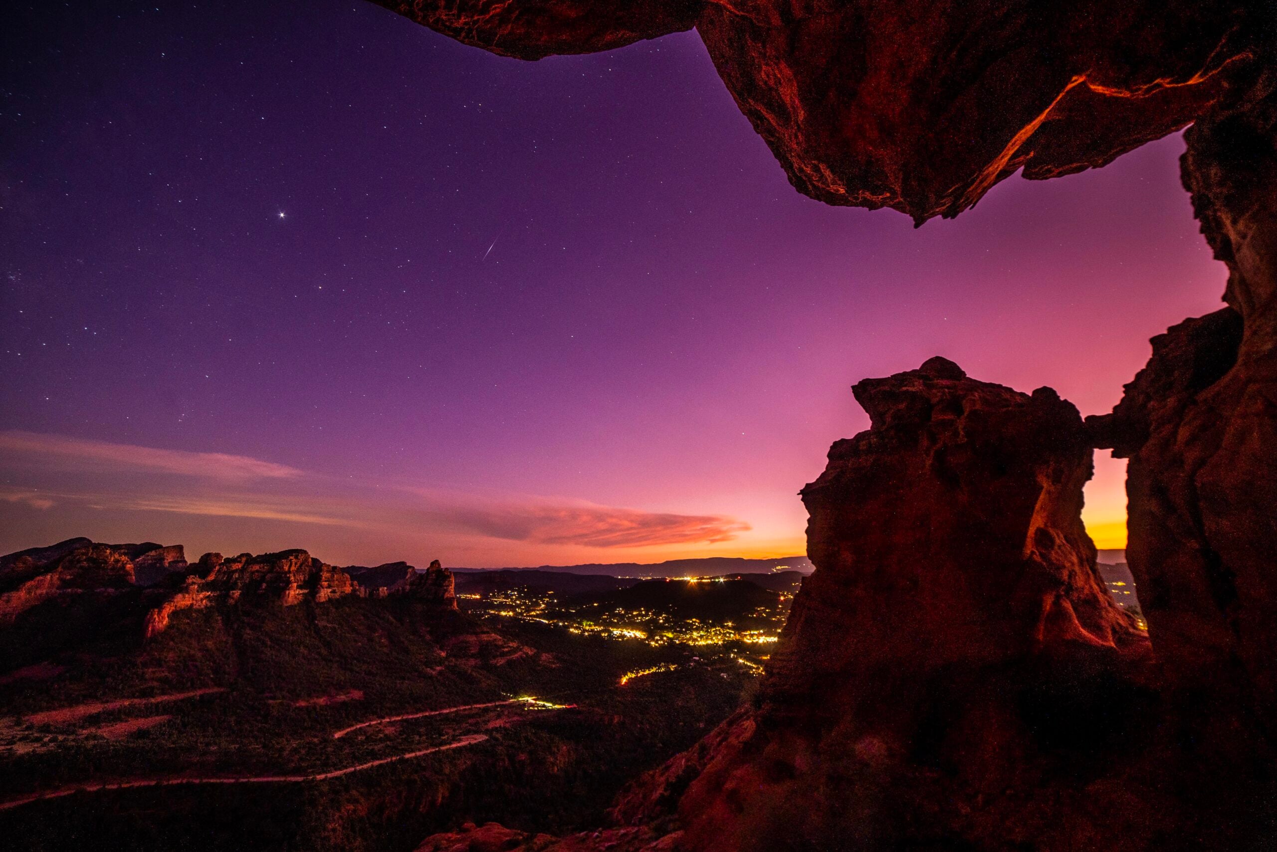 Rocky cliffs frame a glowing purple and orange sunset sky above a distant town with twinkling lights, under a starry night—an unforgettable scene often captured in photos hikers have posted after reaching the summit.