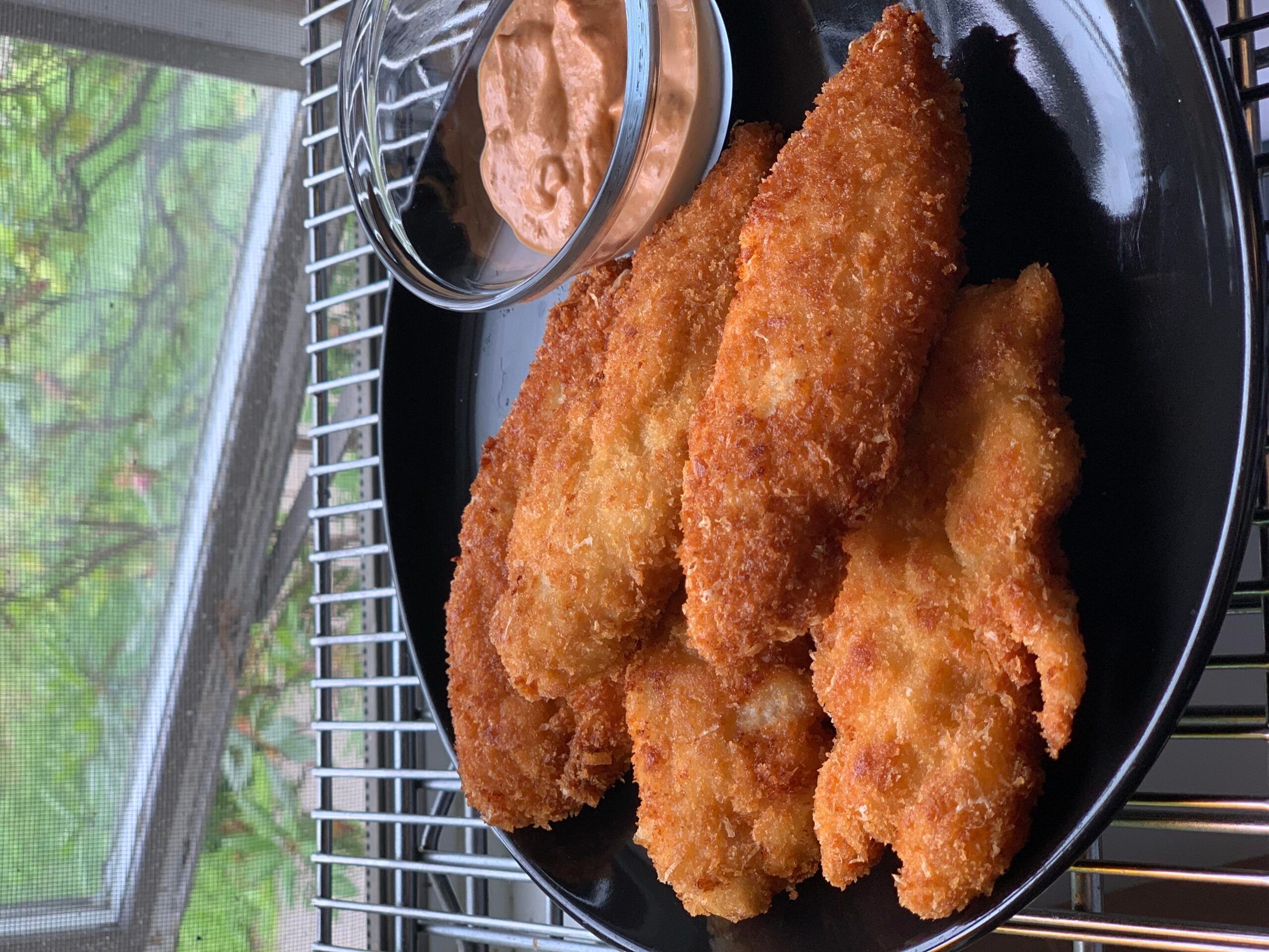 A black plate holds several pieces of crispy fried chicken tenders next to a small glass bowl of dipping sauce, set on a metal rack near a window.